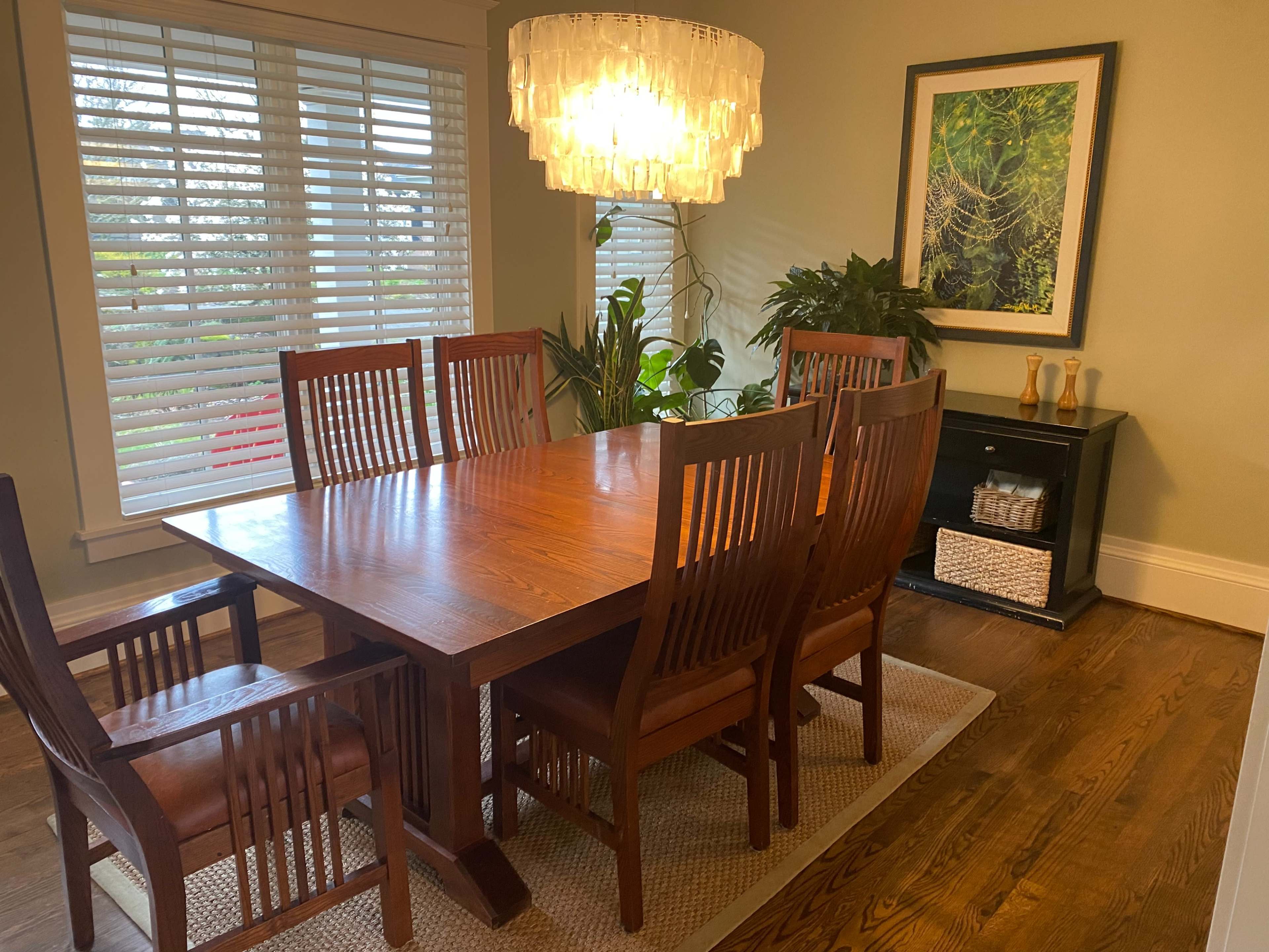 A wooden dining table with eight chairs is set beneath a chandelier, surrounded by large windows with blinds and a wall art piece featuring greenery.