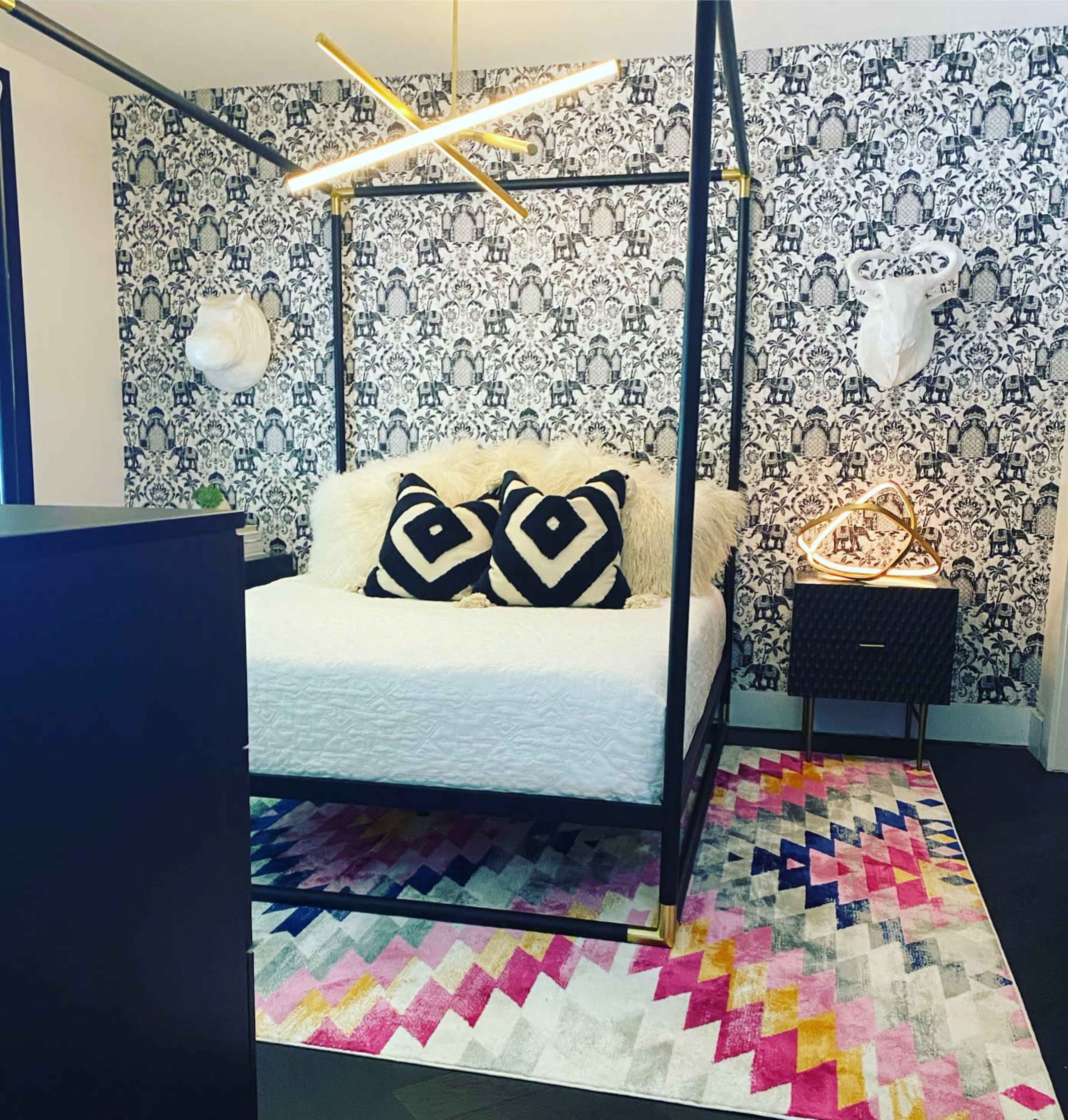 The image shows a modern bedroom featuring a canopy bed with a white bedspread, black and white patterned throw pillows, decorative wall art, and a colorful geometric rug.