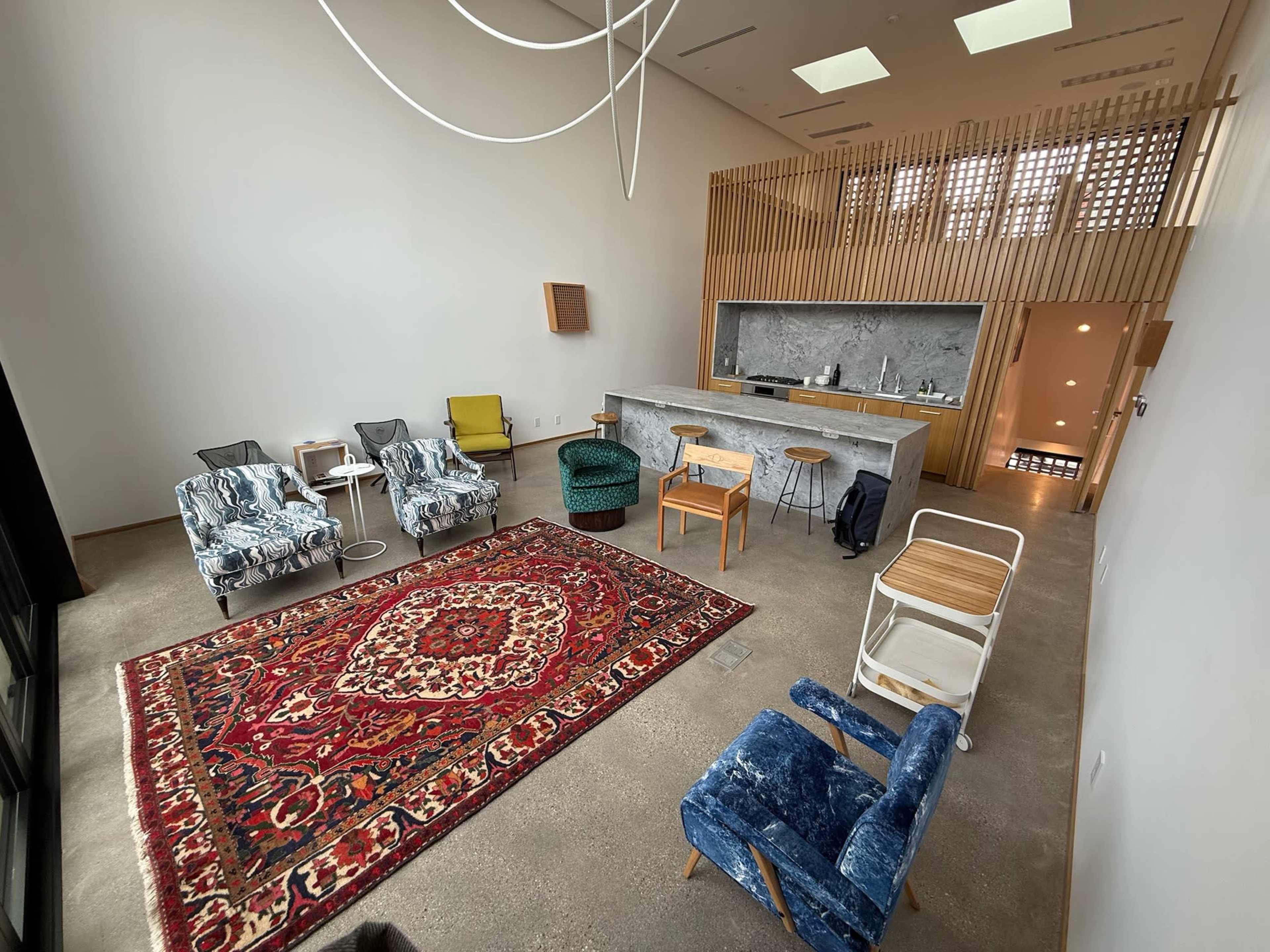 The image depicts a modern interior space featuring a kitchen area with a marble counter, various seating options, and a large decorative rug on a concrete floor.