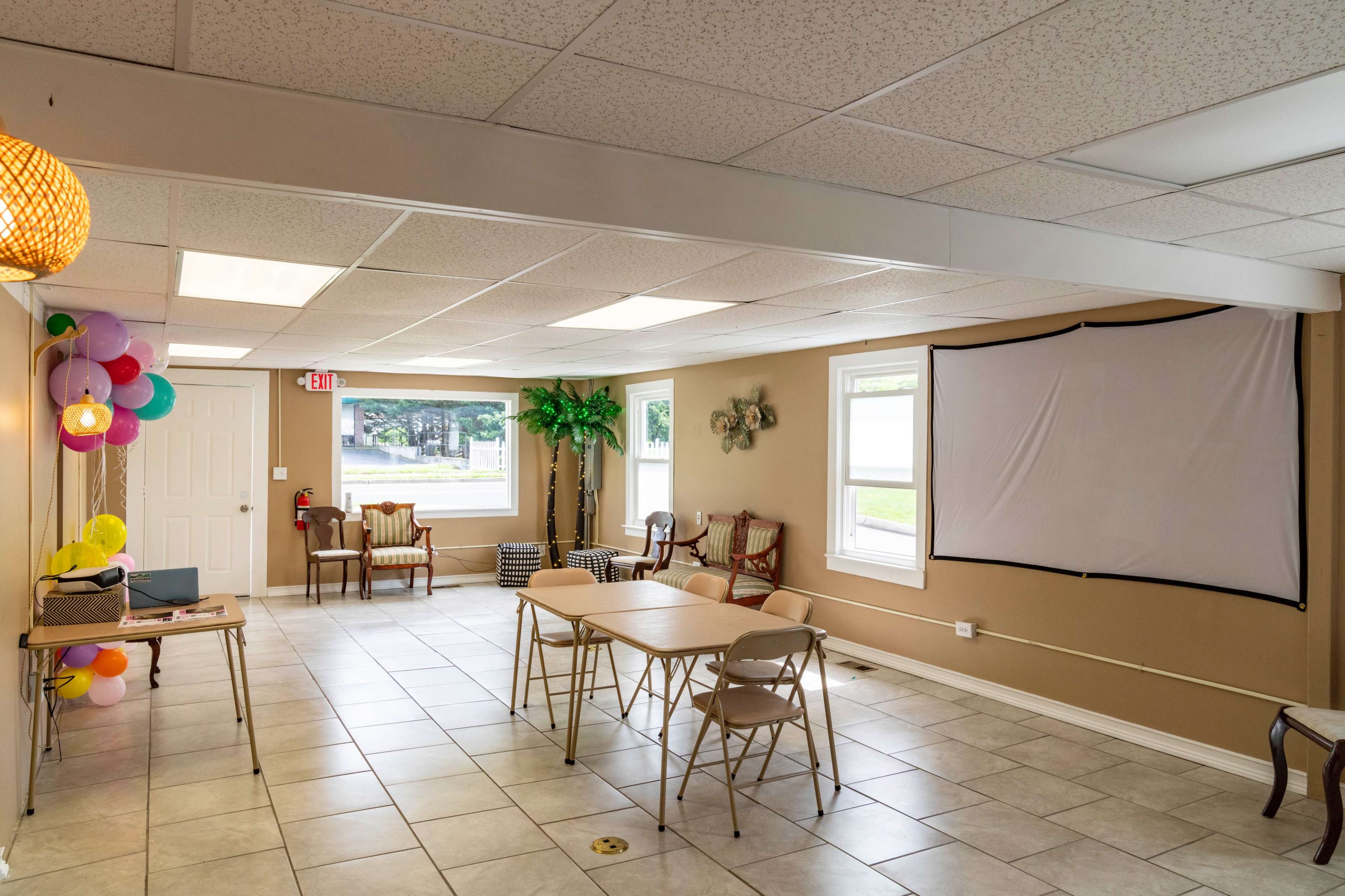 A spacious, well-lit room with tables and chairs arranged for a gathering, featuring a projector screen on one wall.