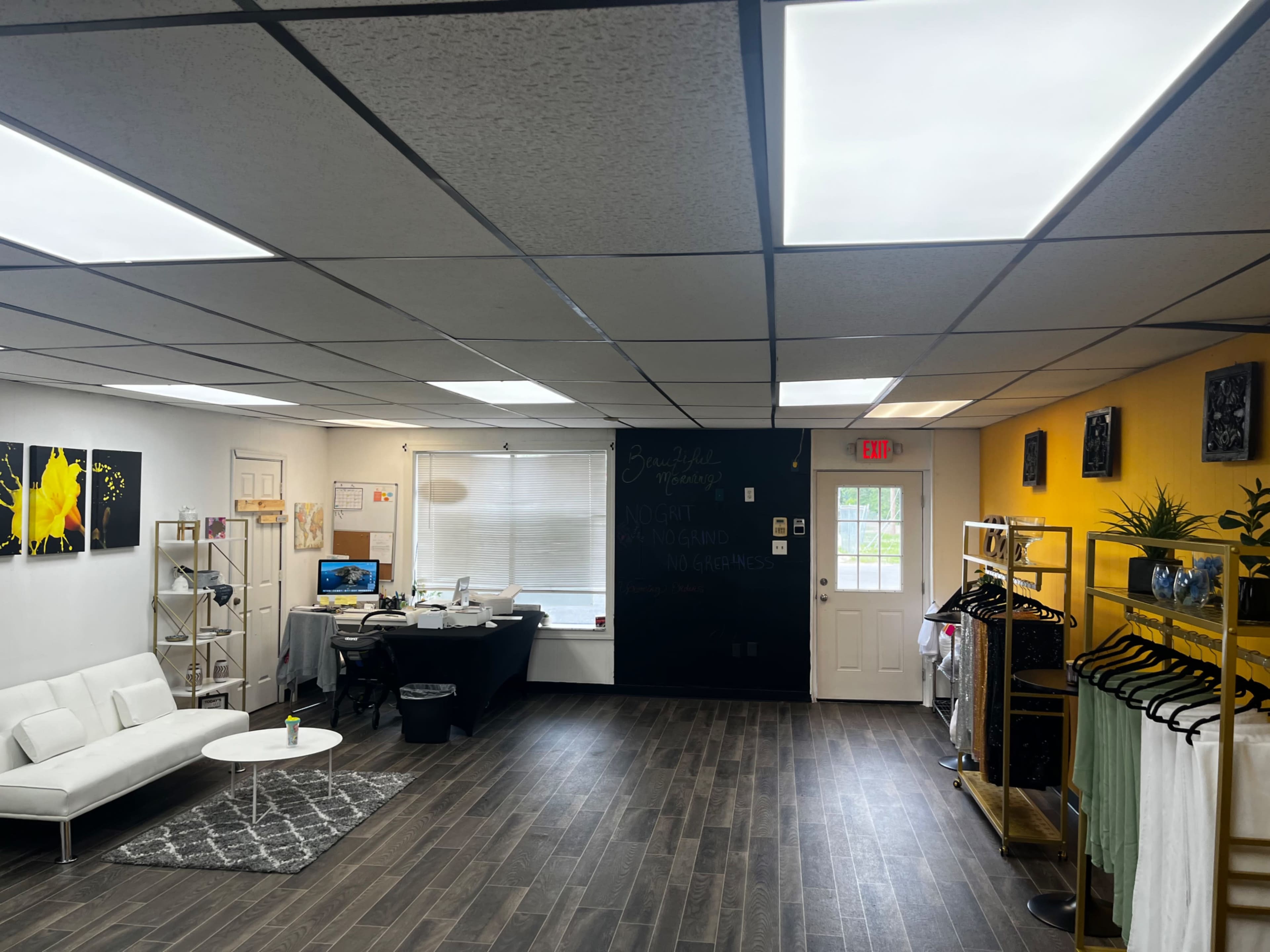 The image shows a bright, modern interior of a retail space with a white sofa, a desk with a computer, and clothing racks against the walls.