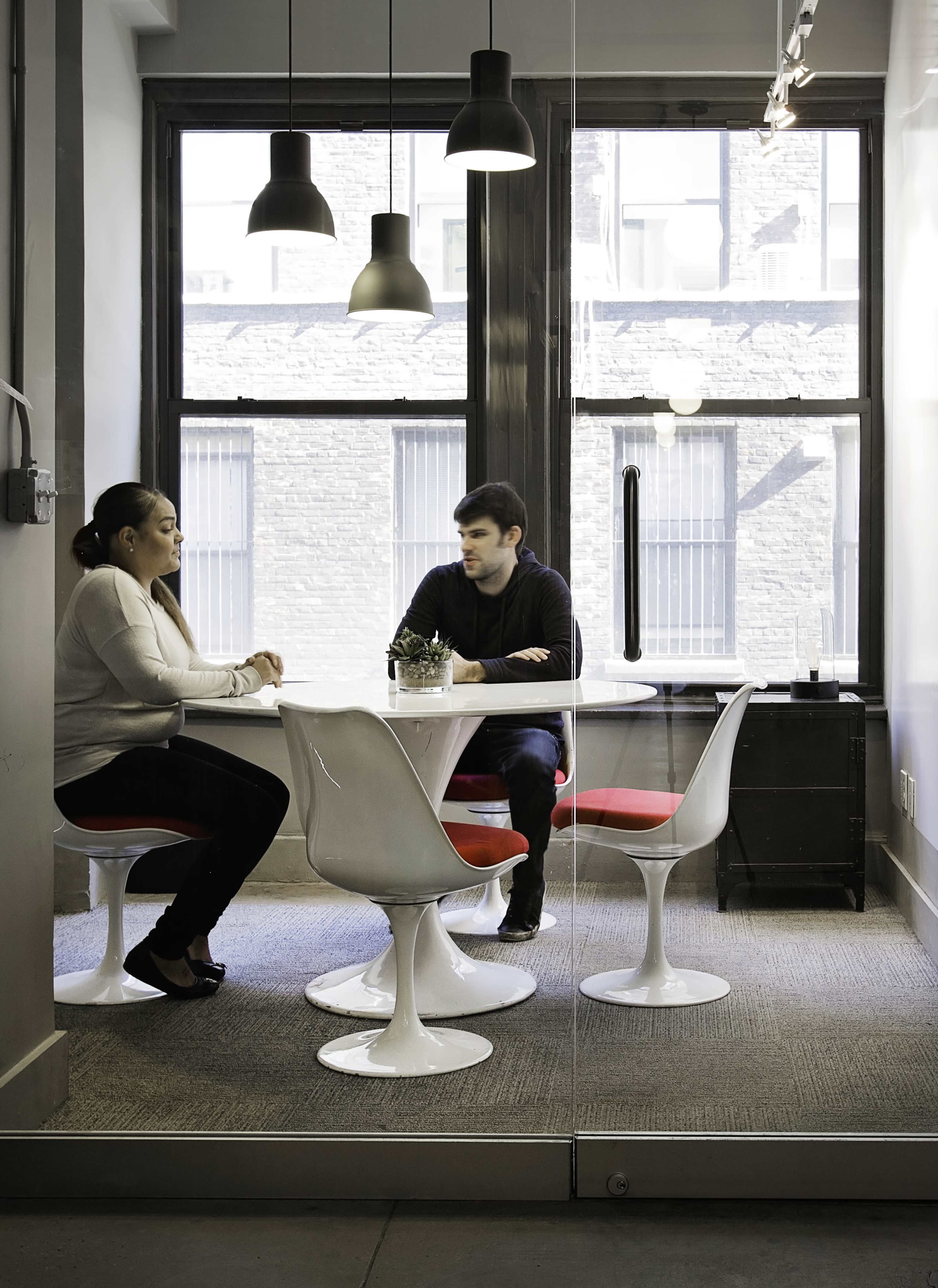 Two people are seated at a round table in a modern office space with large windows and pendant lighting.