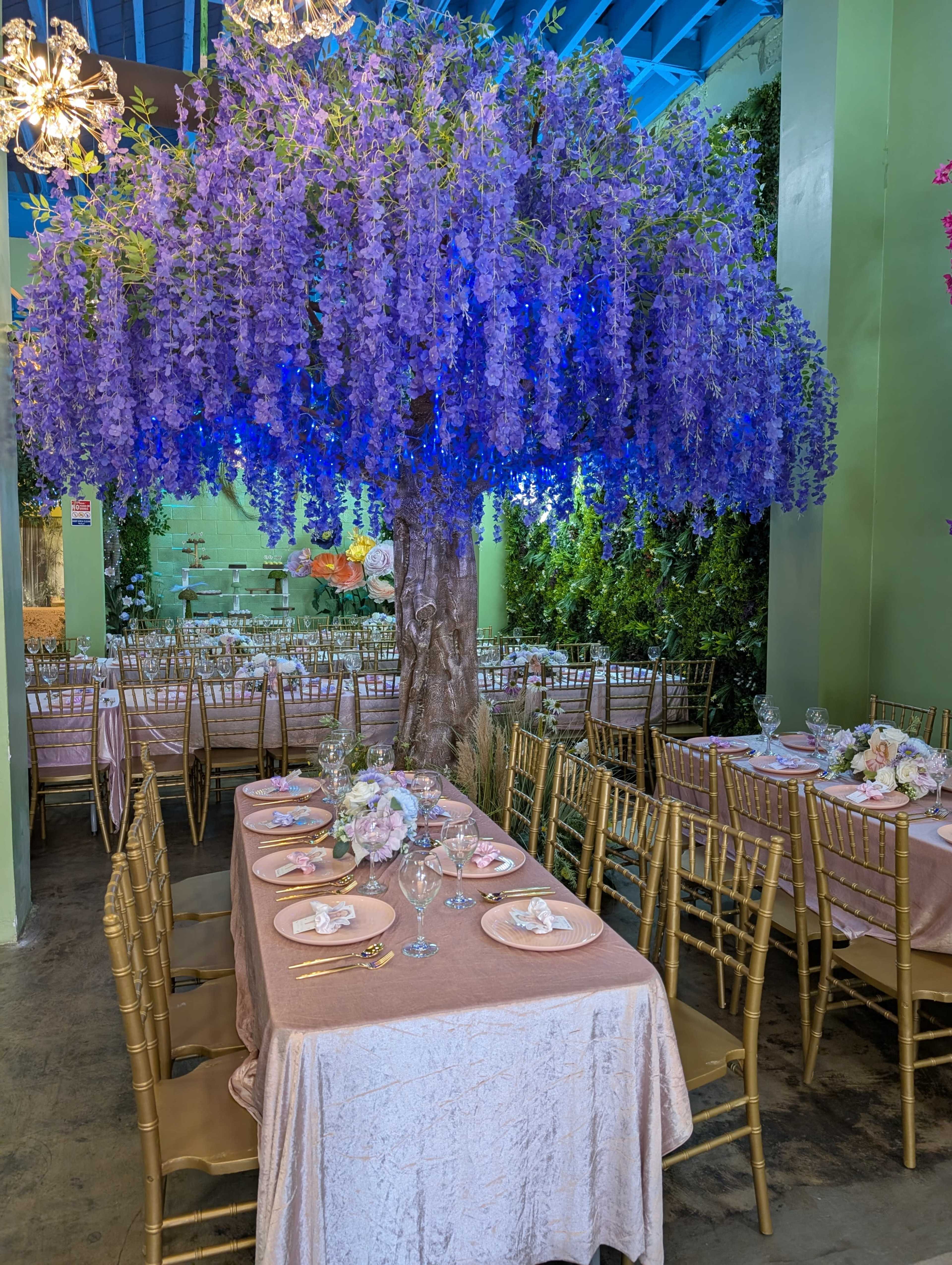The image shows a dining area adorned with a large artificial floral tree featuring purple flowers, tables set with plates and glassware, and gold chairs.