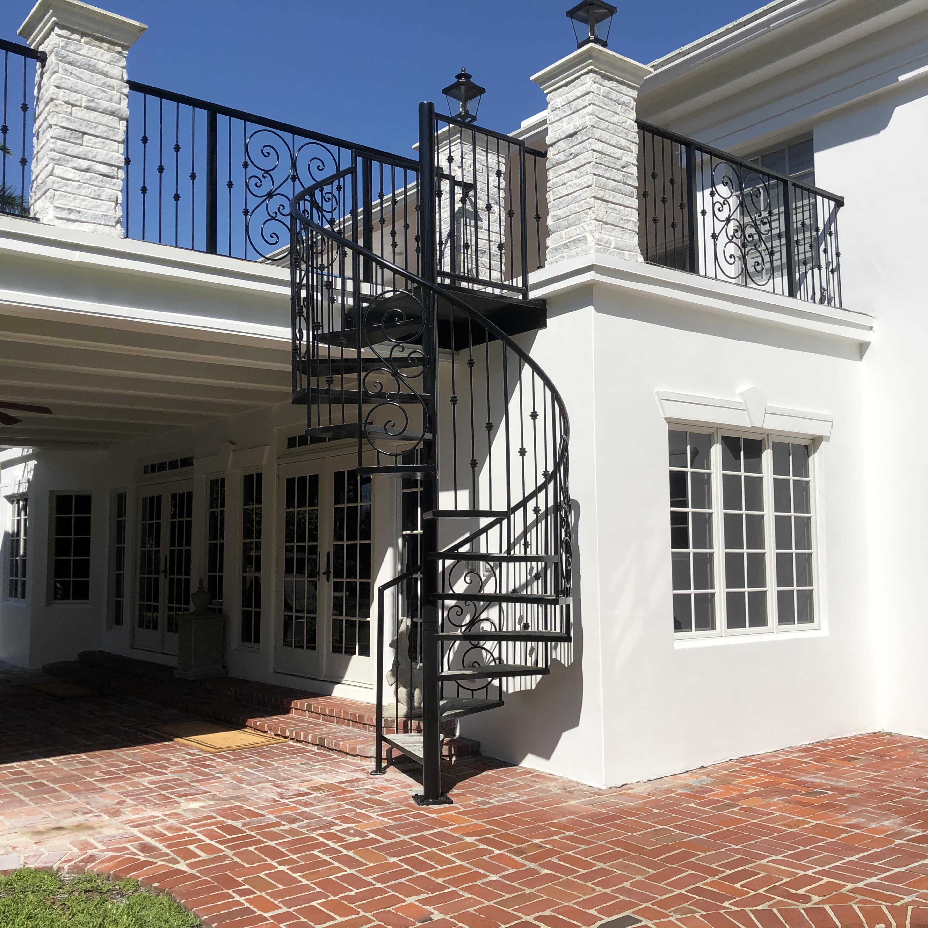 A black spiral staircase leads from a patio to a balcony of a white building with large windows.