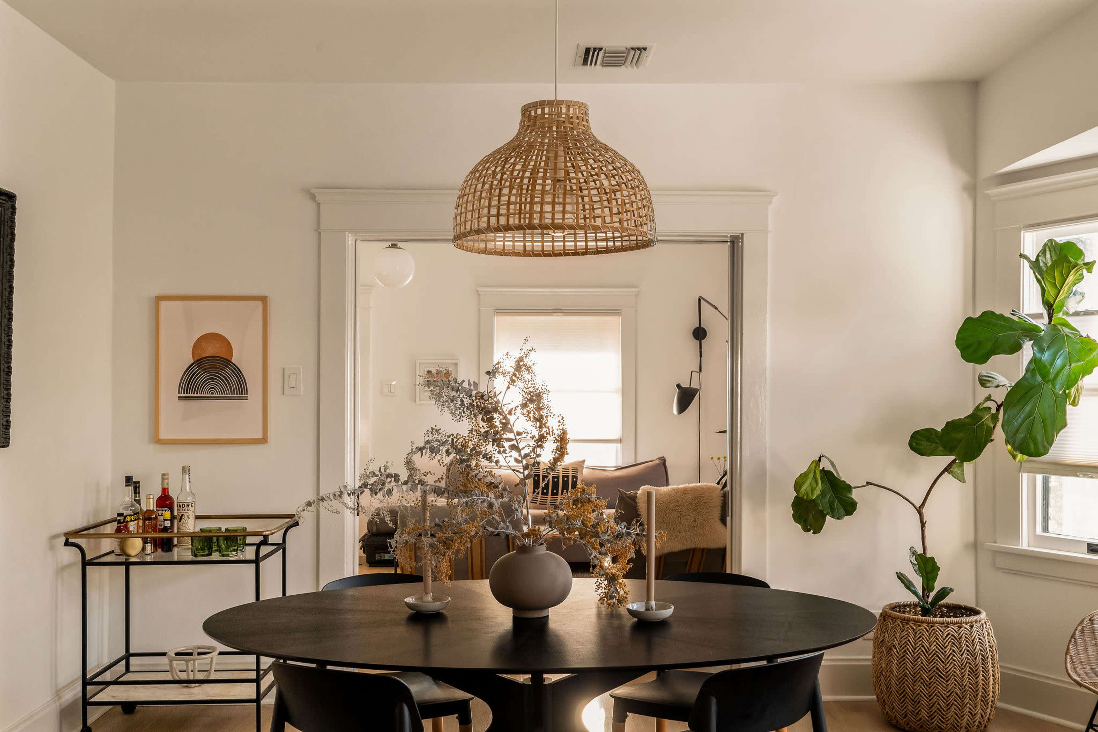 A round black dining table is set in a well-lit room featuring a rattan pendant light overhead and a decorative arrangement of dried branches.