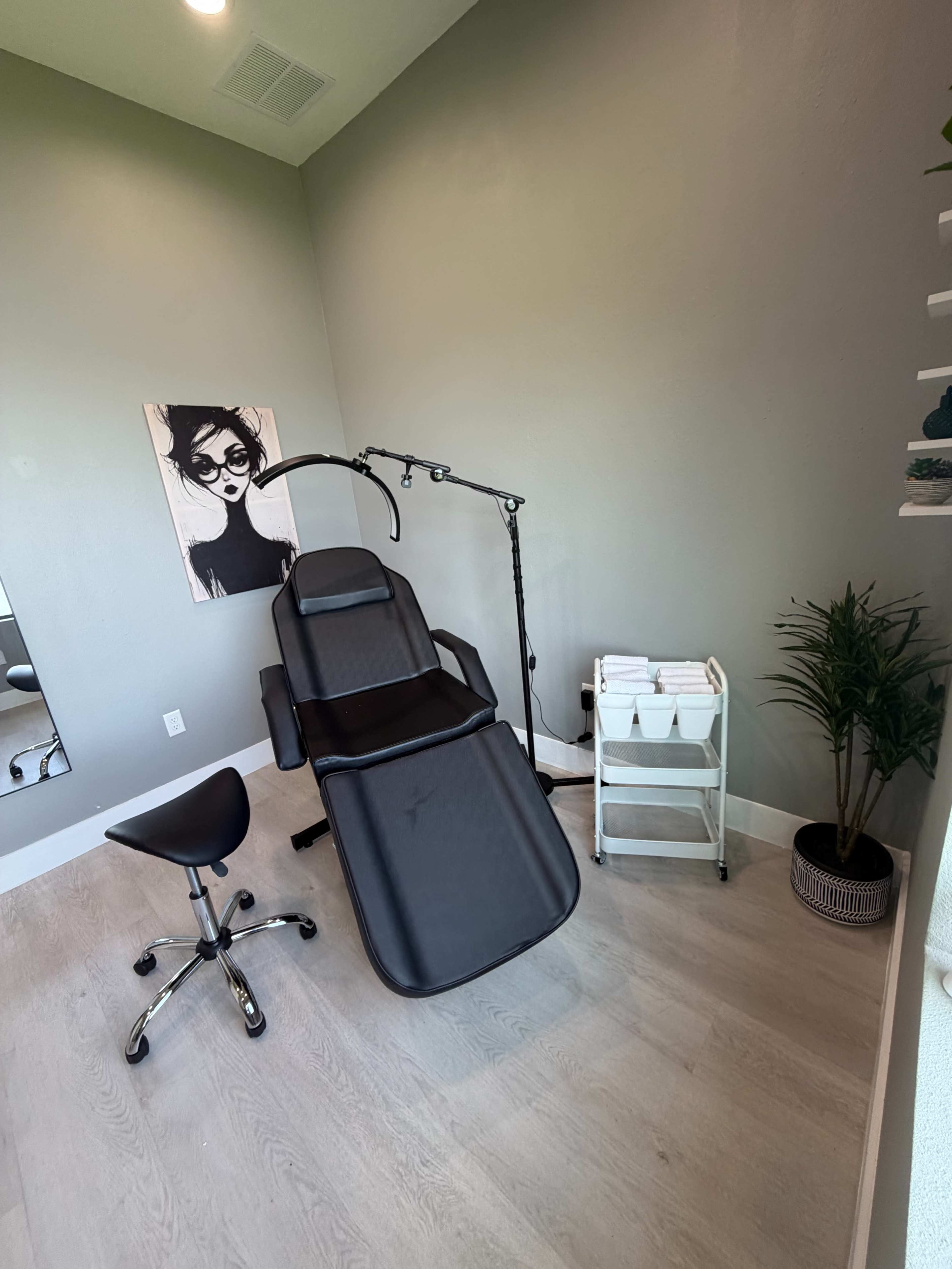 The image shows a small, well-lit beauty treatment room featuring a black treatment chair, a stool, a cart with towels, and a wall-mounted lamp, along with an artistic portrait on the wall.