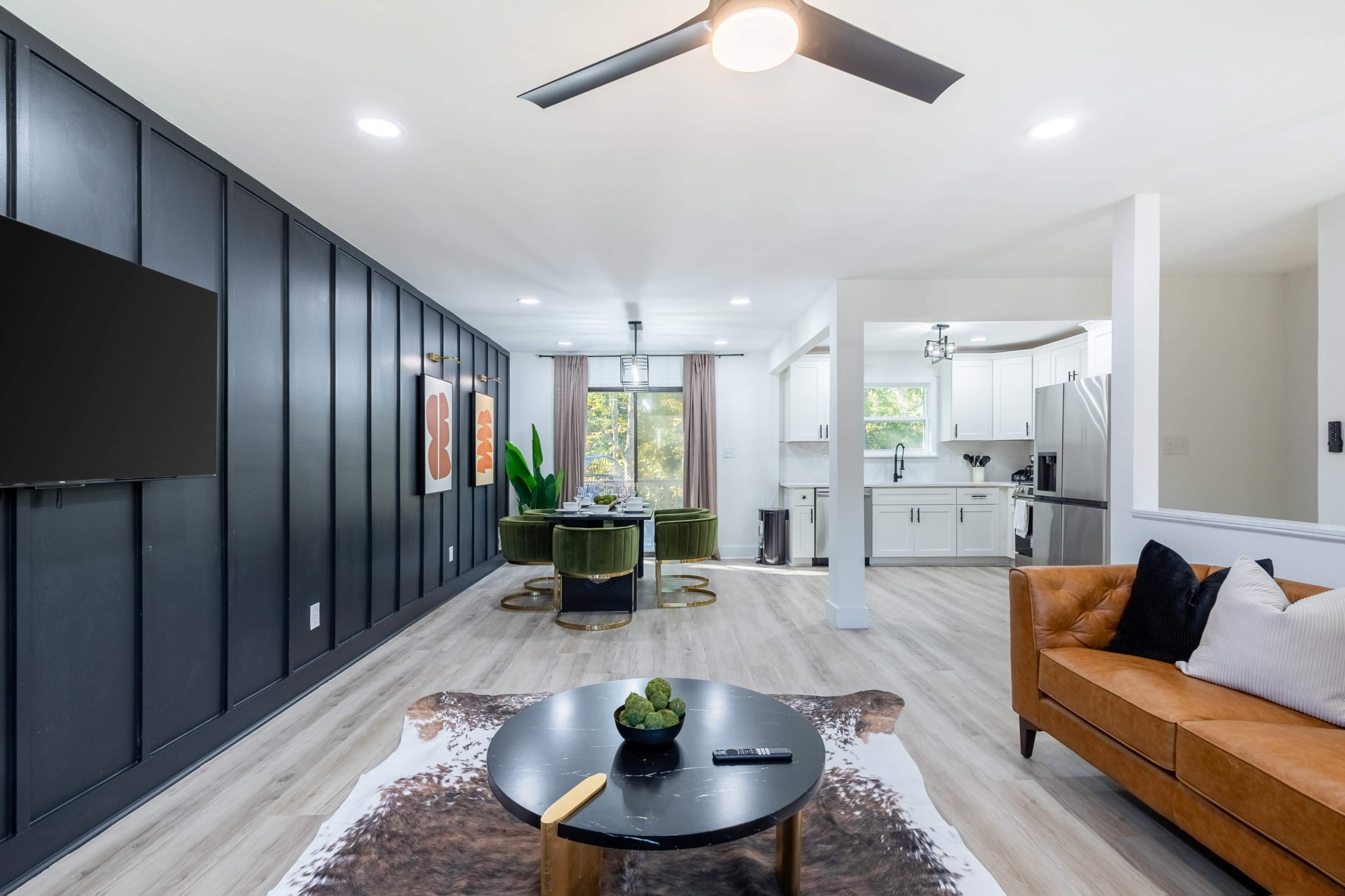 The image shows a modern living space featuring a brown leather sofa, a black accent wall with a TV, and a green velvet seating area adjacent to a kitchen.
