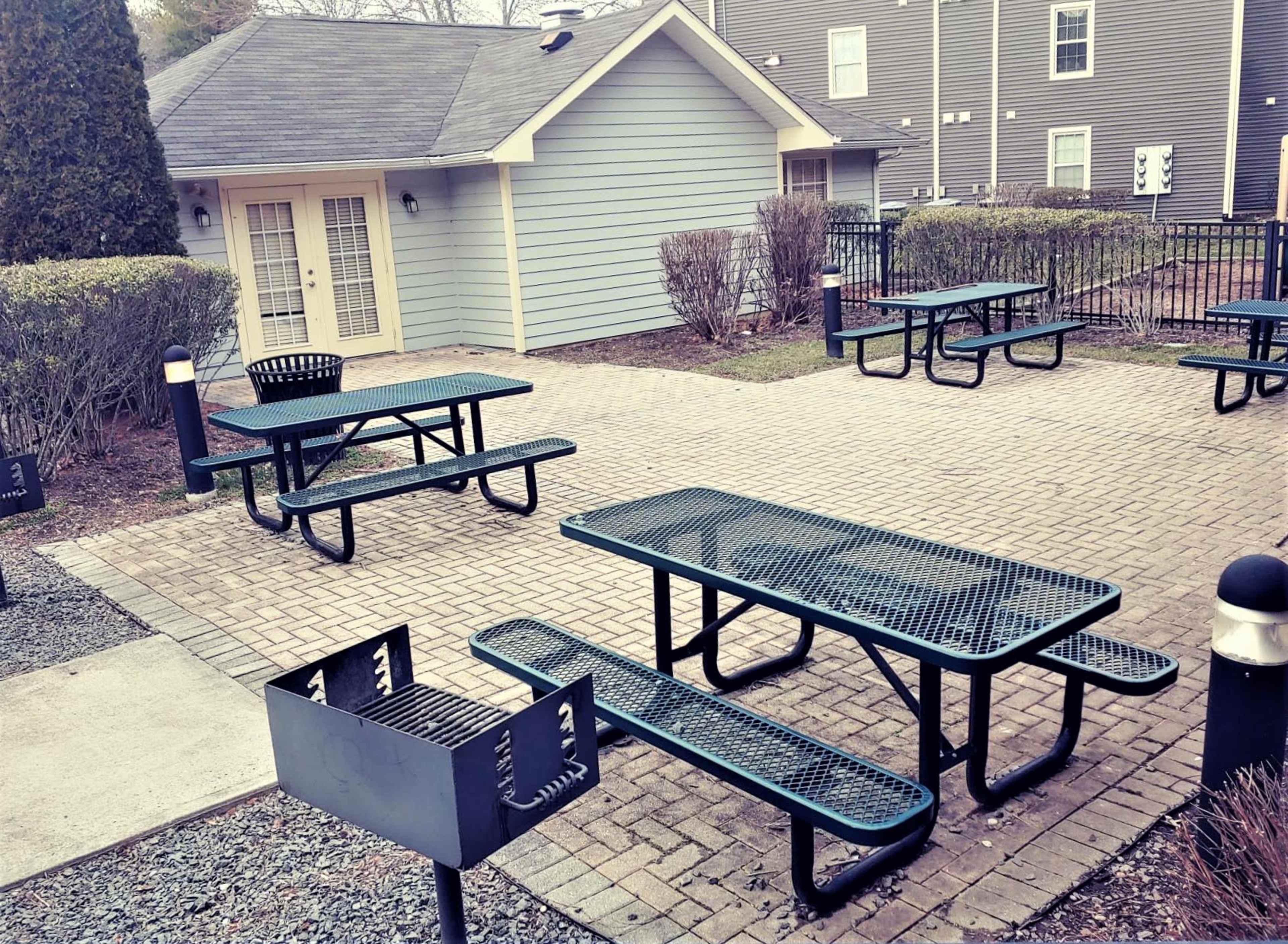 A patio area with several green picnic tables and grills surrounded by low shrubs and a gray building in the background.