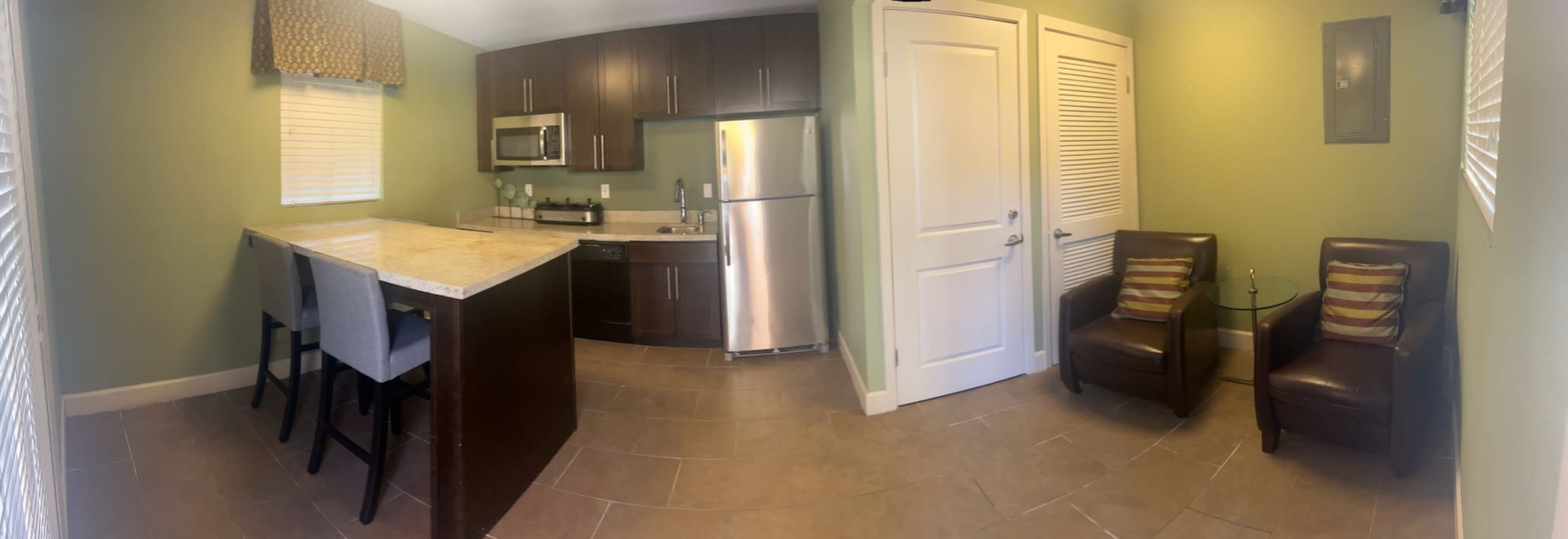 A modern kitchen and living area featuring a dark wood island with two bar stools, stainless steel appliances, and two brown chairs with cushions.