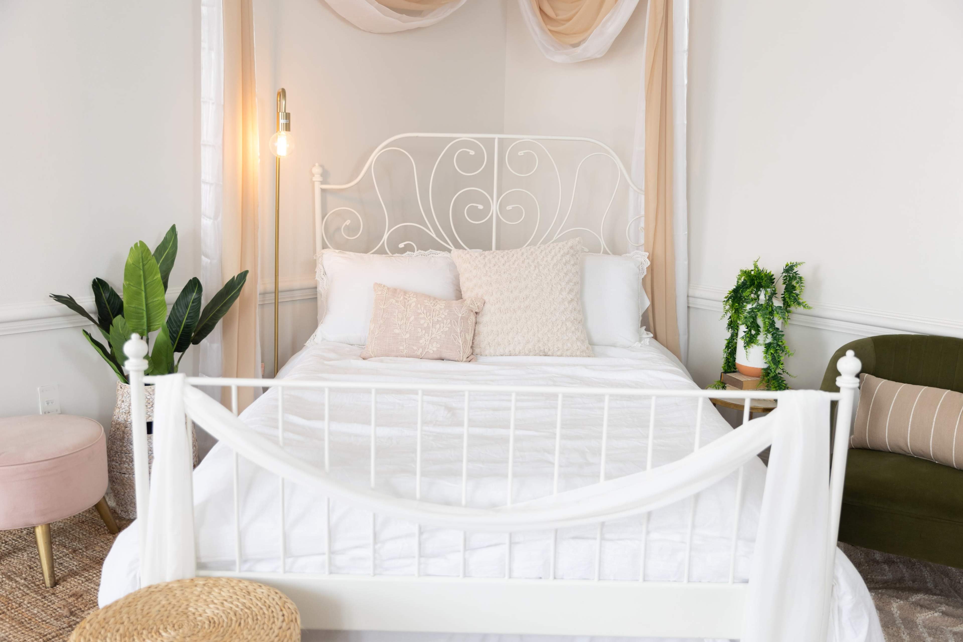 A well-lit bedroom with a white wrought-iron bed, adorned with decorative pillows, and surrounded by plants and a green armchair.