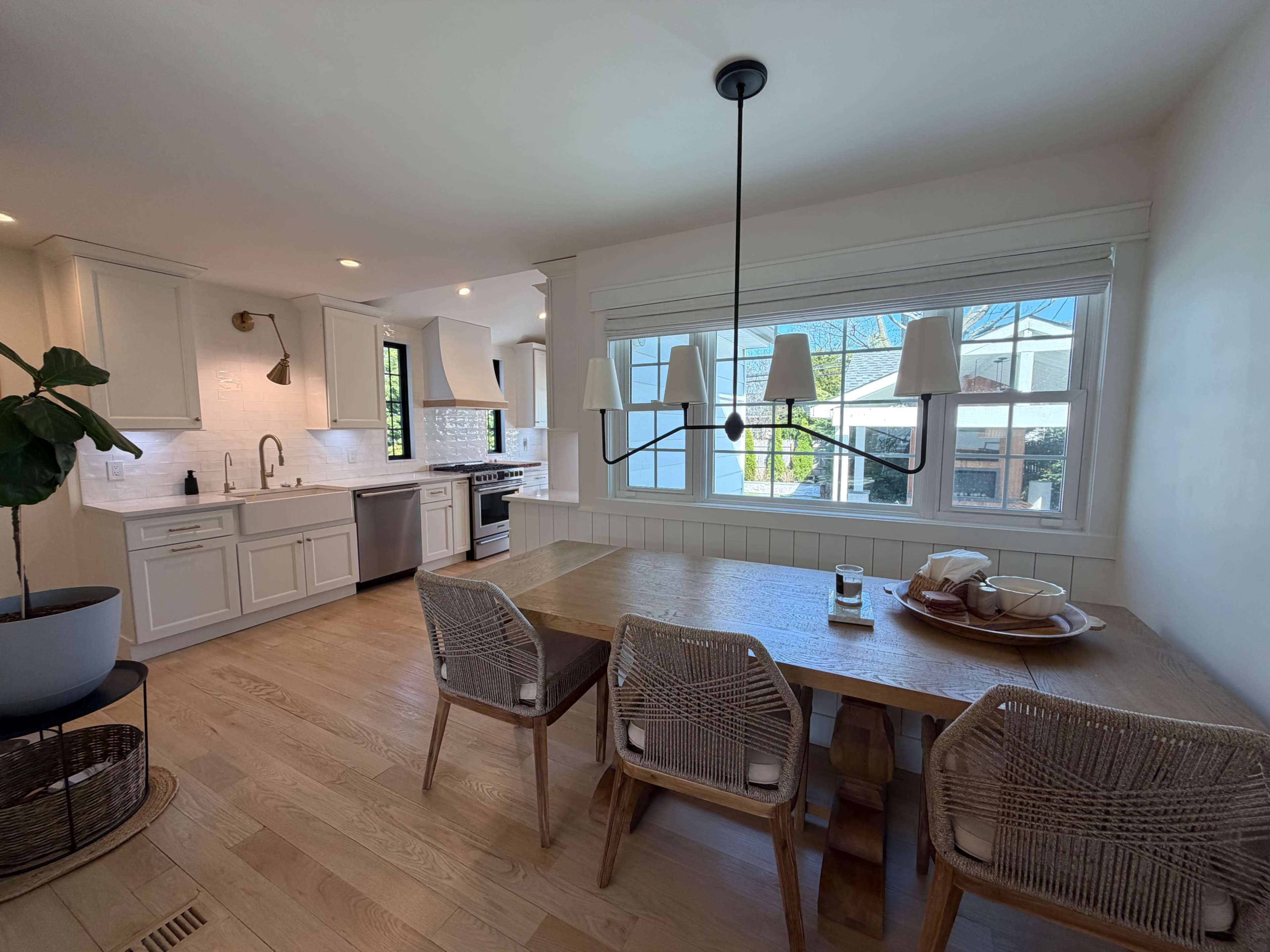 A modern kitchen features white cabinetry, stainless steel appliances, and a wooden dining table surrounded by woven chairs, with large windows providing natural light.