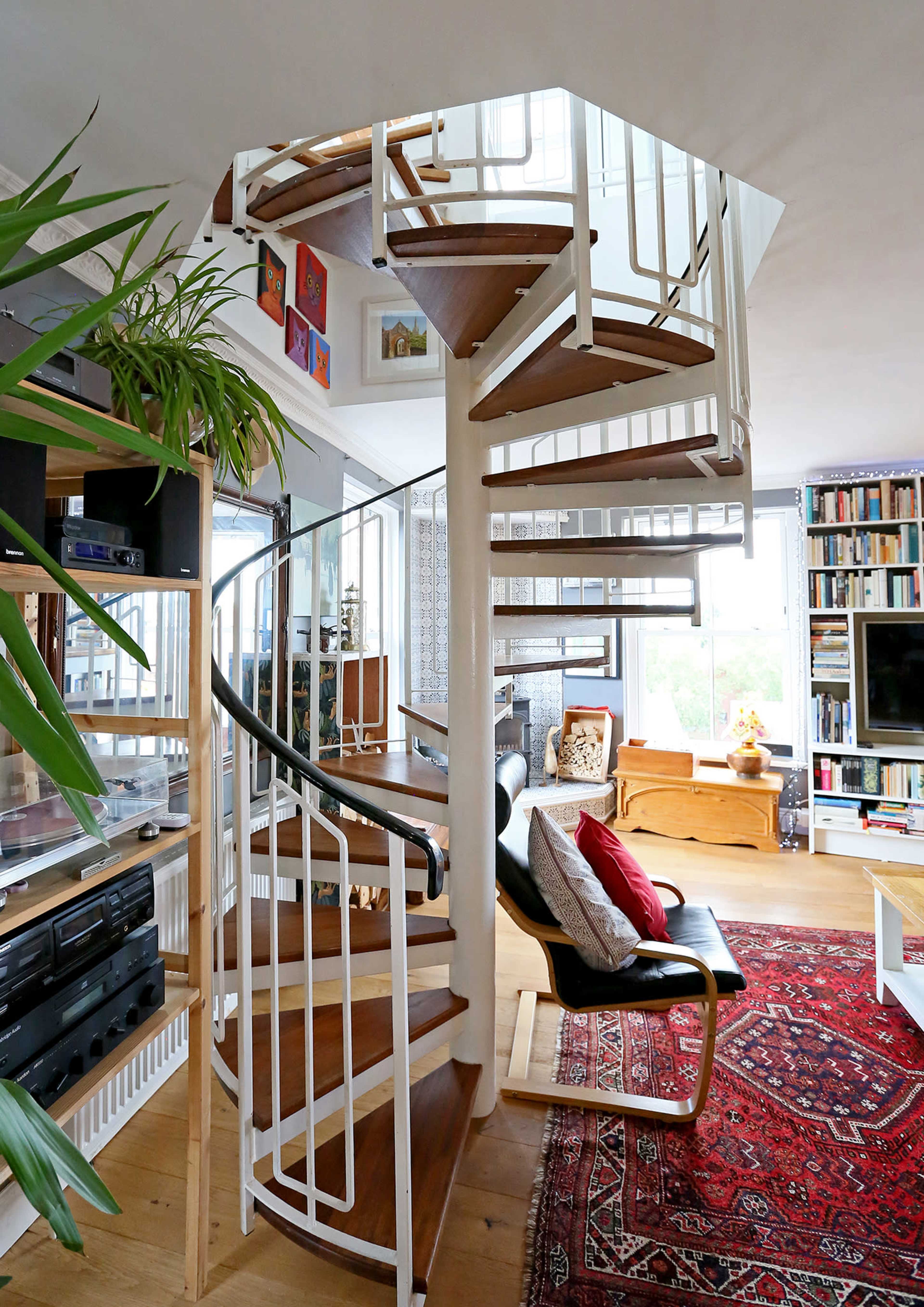 A spiral staircase with wooden steps and a glass railing is situated in a well-furnished living area featuring bookshelves and decorative elements.
