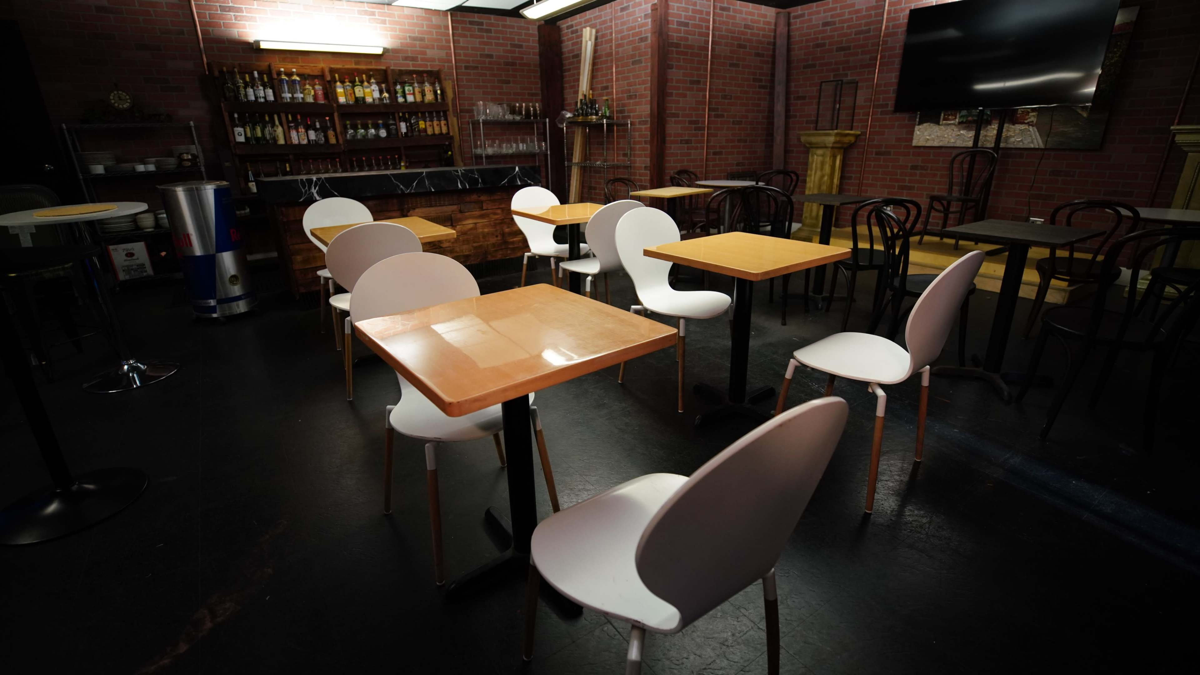 An empty restaurant interior with several tables and chairs arranged on a dark floor, surrounded by brick walls and a bar area in the background.