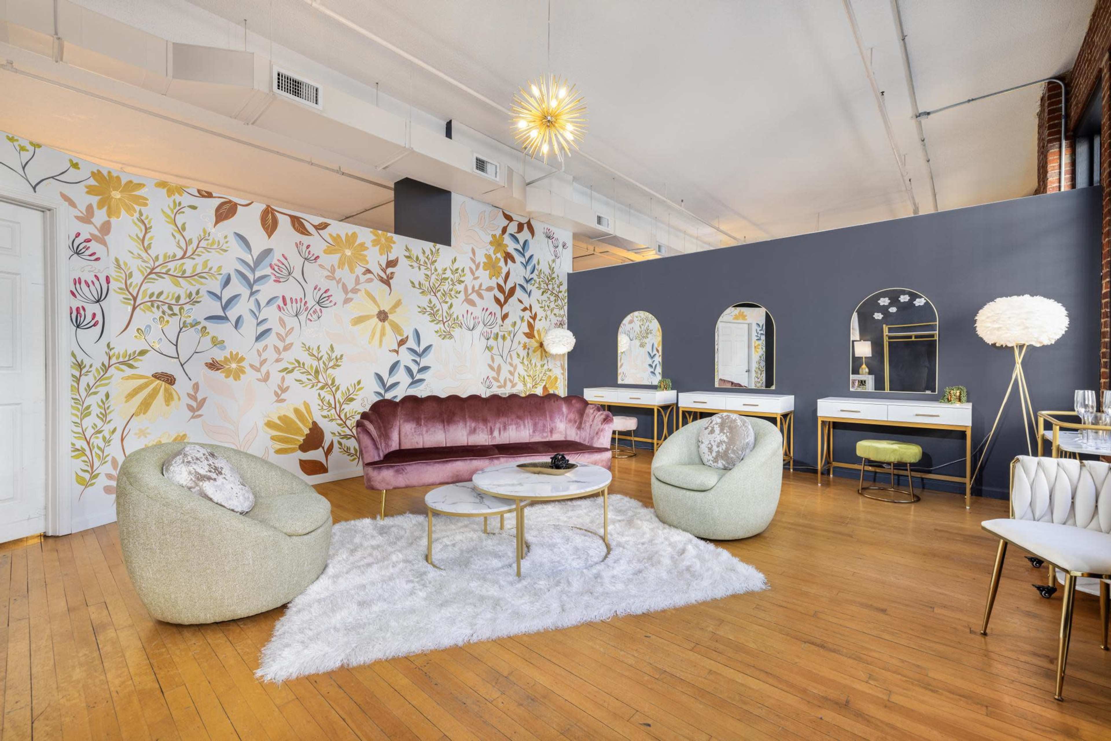 The room features a blend of floral wallpaper, a pink velvet sofa, two light-colored chairs, a round coffee table, and several mirrors set against a dark wall.