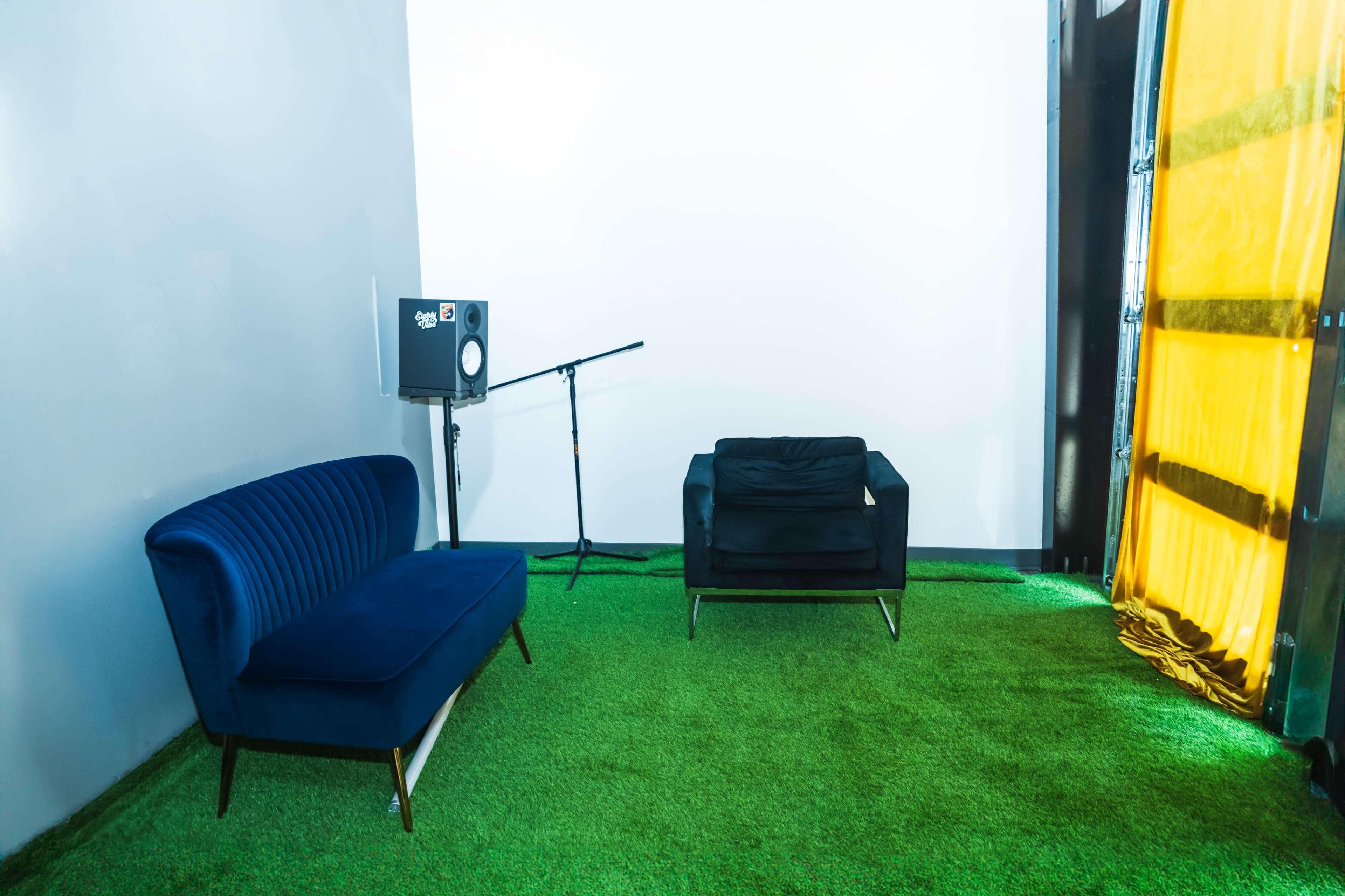 The image shows a simple room with artificial green grass flooring, featuring a blue sofa, a black armchair, a microphone on a stand, and a speaker next to a bright yellow curtain.