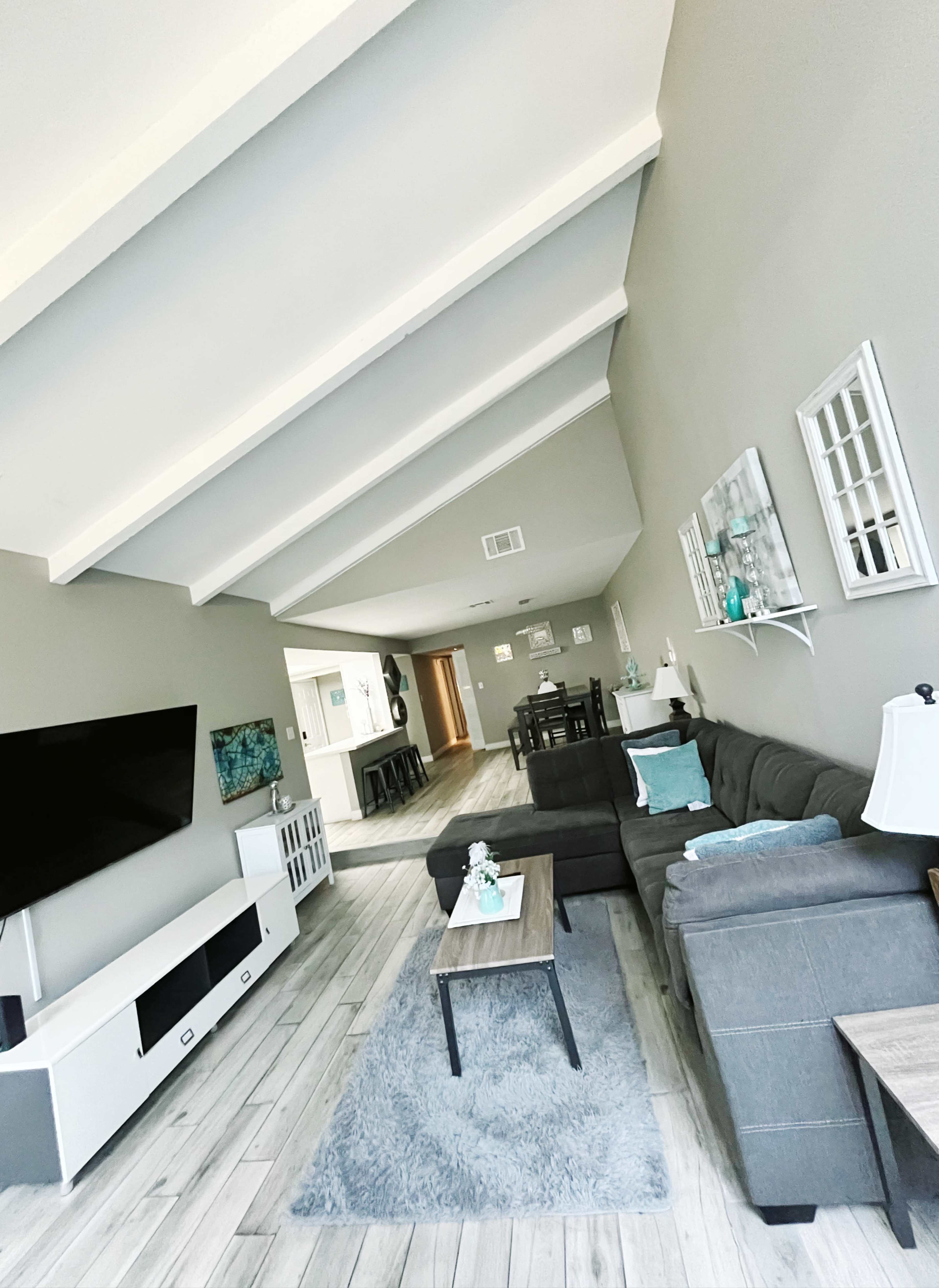 A spacious living room with a gray sectional sofa, a coffee table, and a television on a white media console, featuring vaulted ceilings and light wooden flooring.