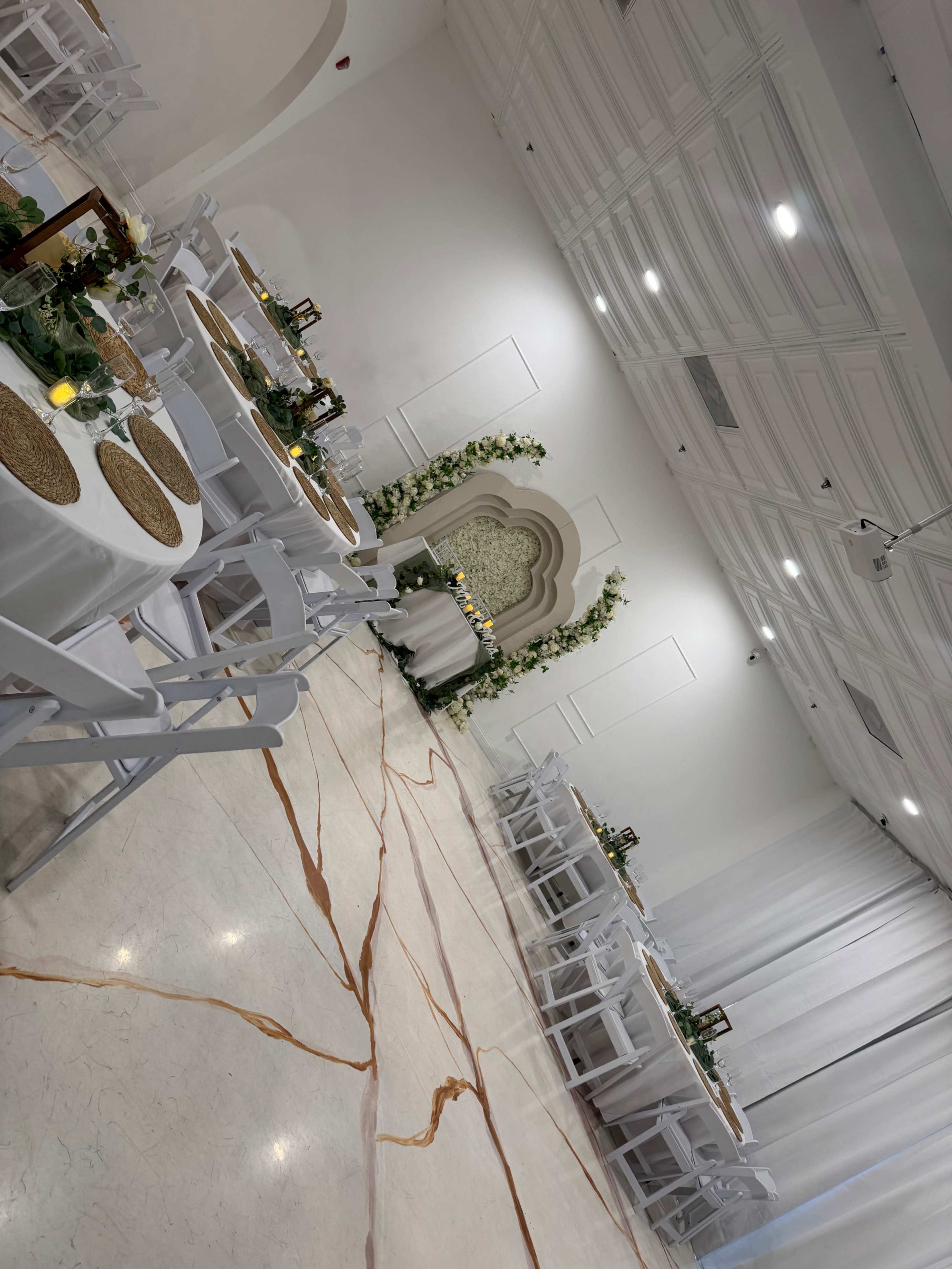 The image shows a elegantly arranged event space with white tables set with woven placemats and decorated with greenery, candles, and floral accents.