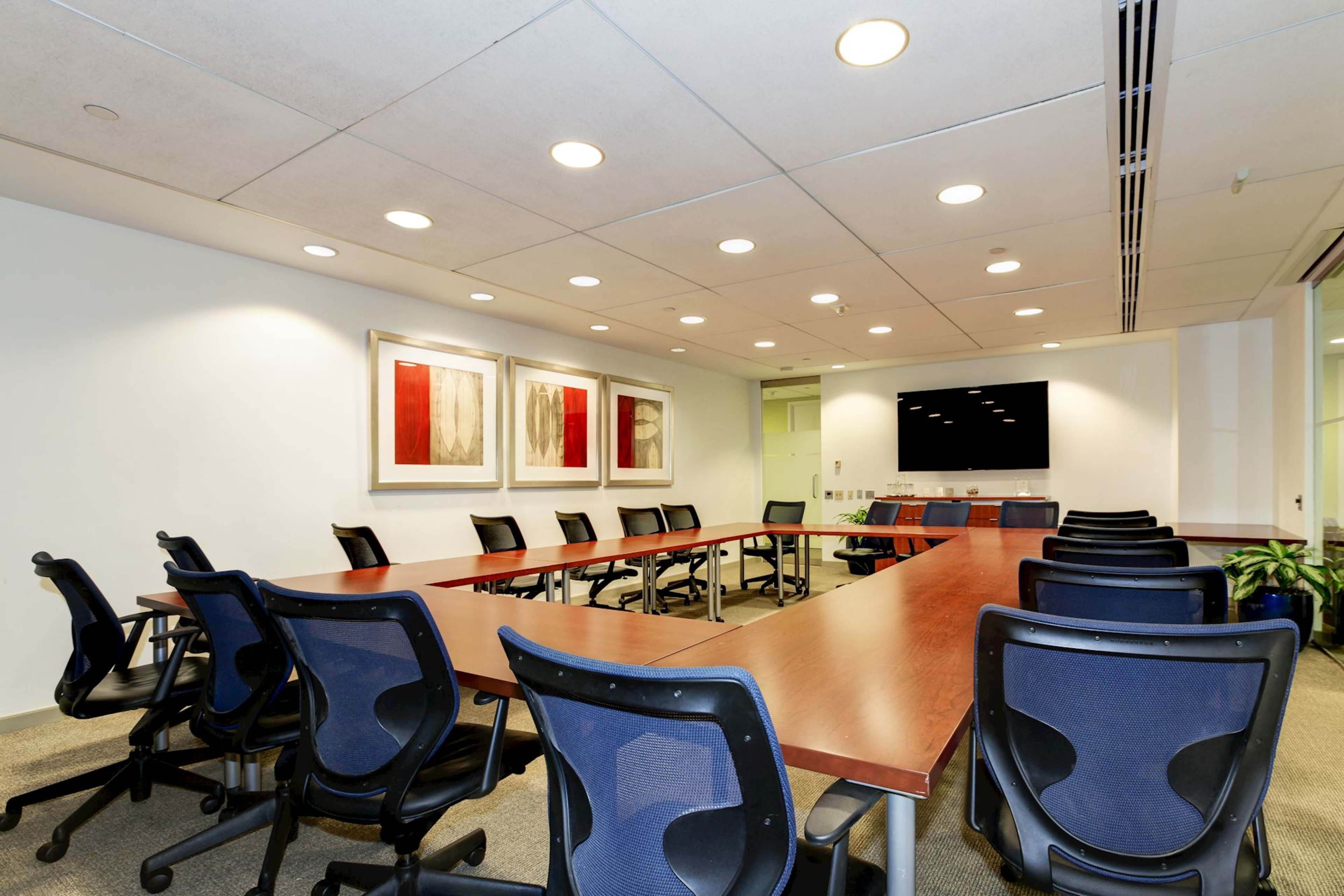 A modern conference room with a long table surrounded by black chairs, featuring two abstract paintings on the wall and a large screen at one end.