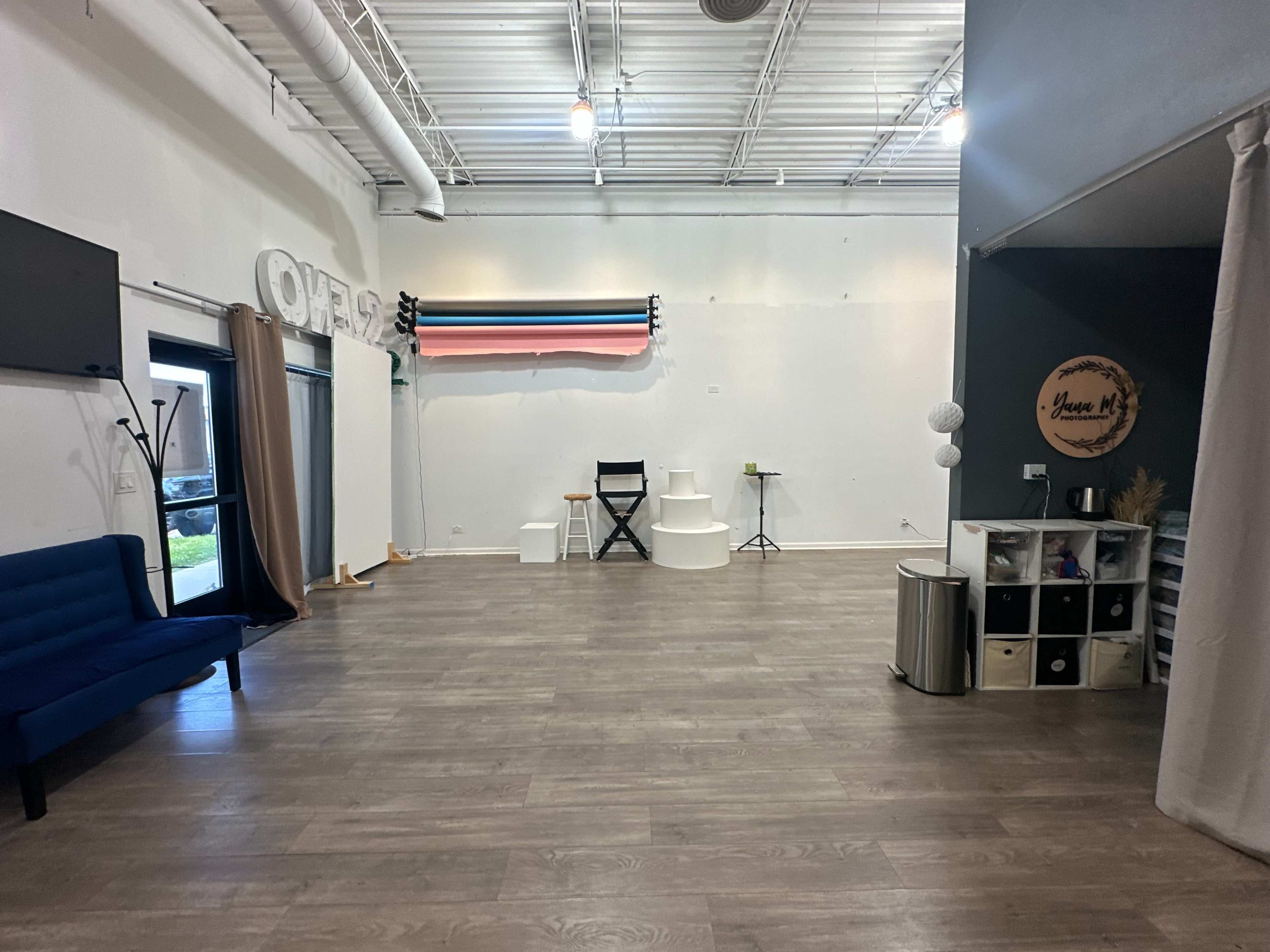 The image shows a spacious interior with hardwood flooring, a blue couch, a black director's chair, a backdrop with colored stripes, and various decorative elements along the walls.