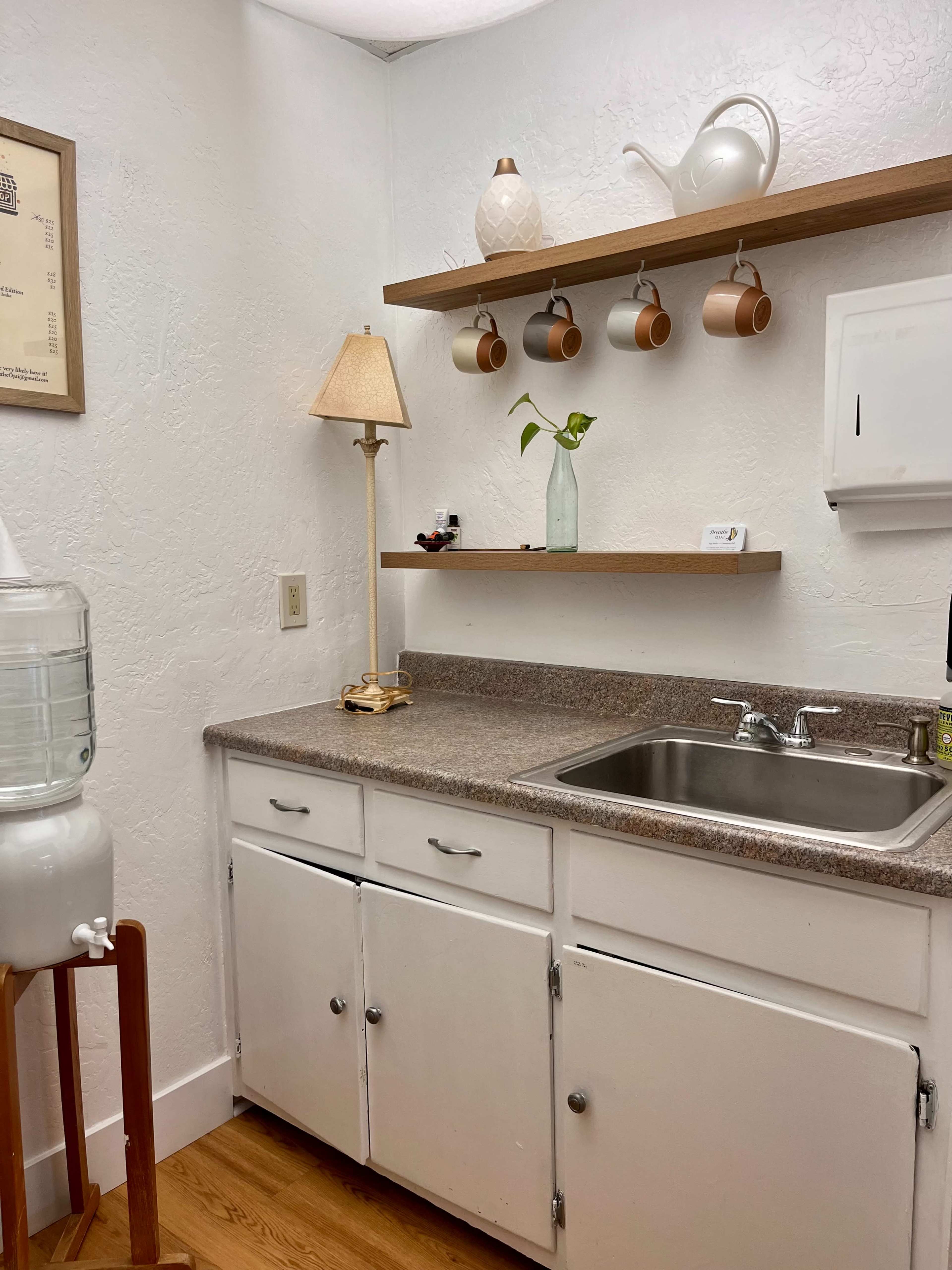 A small kitchen corner with a sink, a water cooler, a lamp, a shelf holding decorative items, and several hanging coffee mugs.