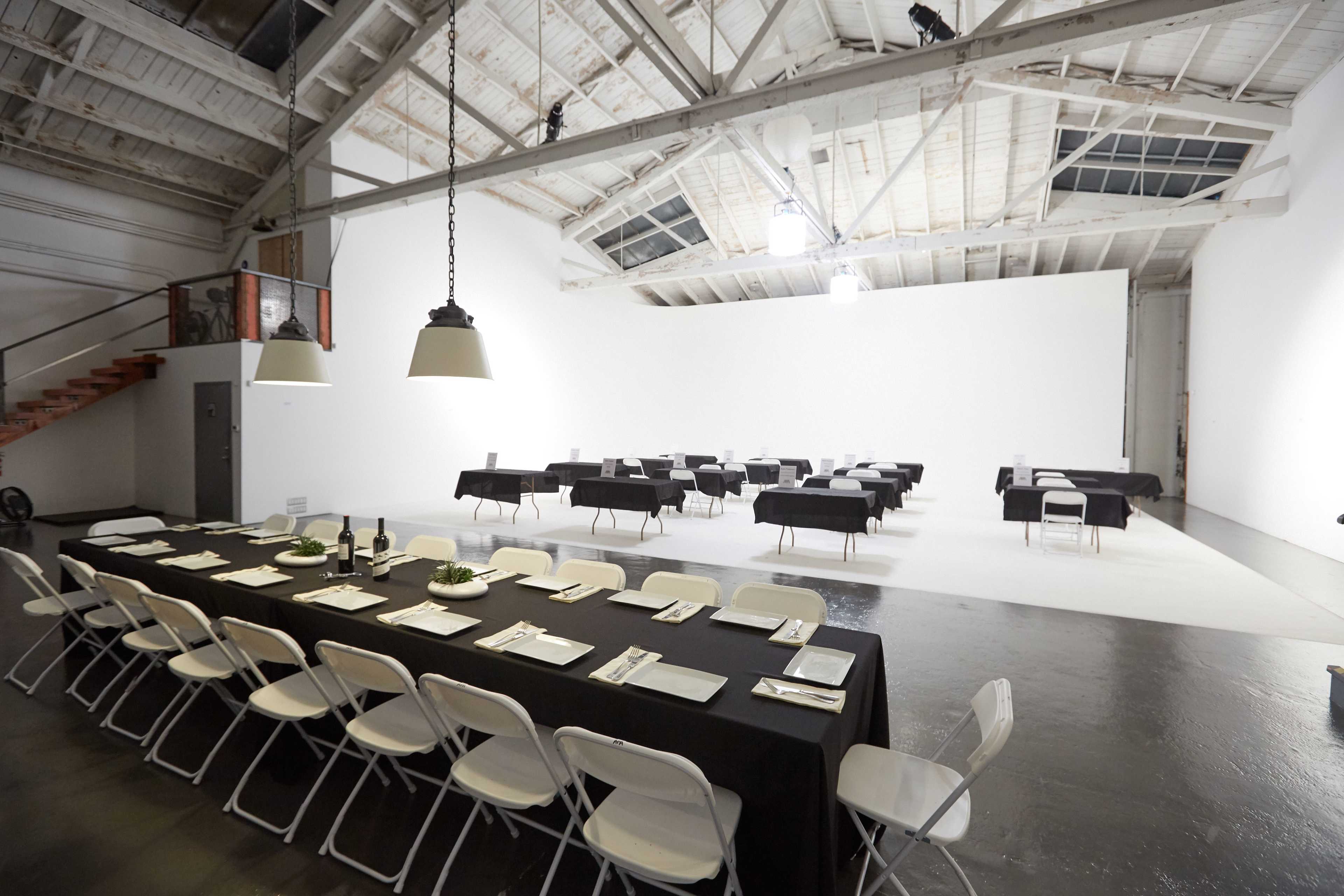 The image shows a spacious, well-lit room with two sets of tables covered in black tablecloths, arranged for a dining or event setup.