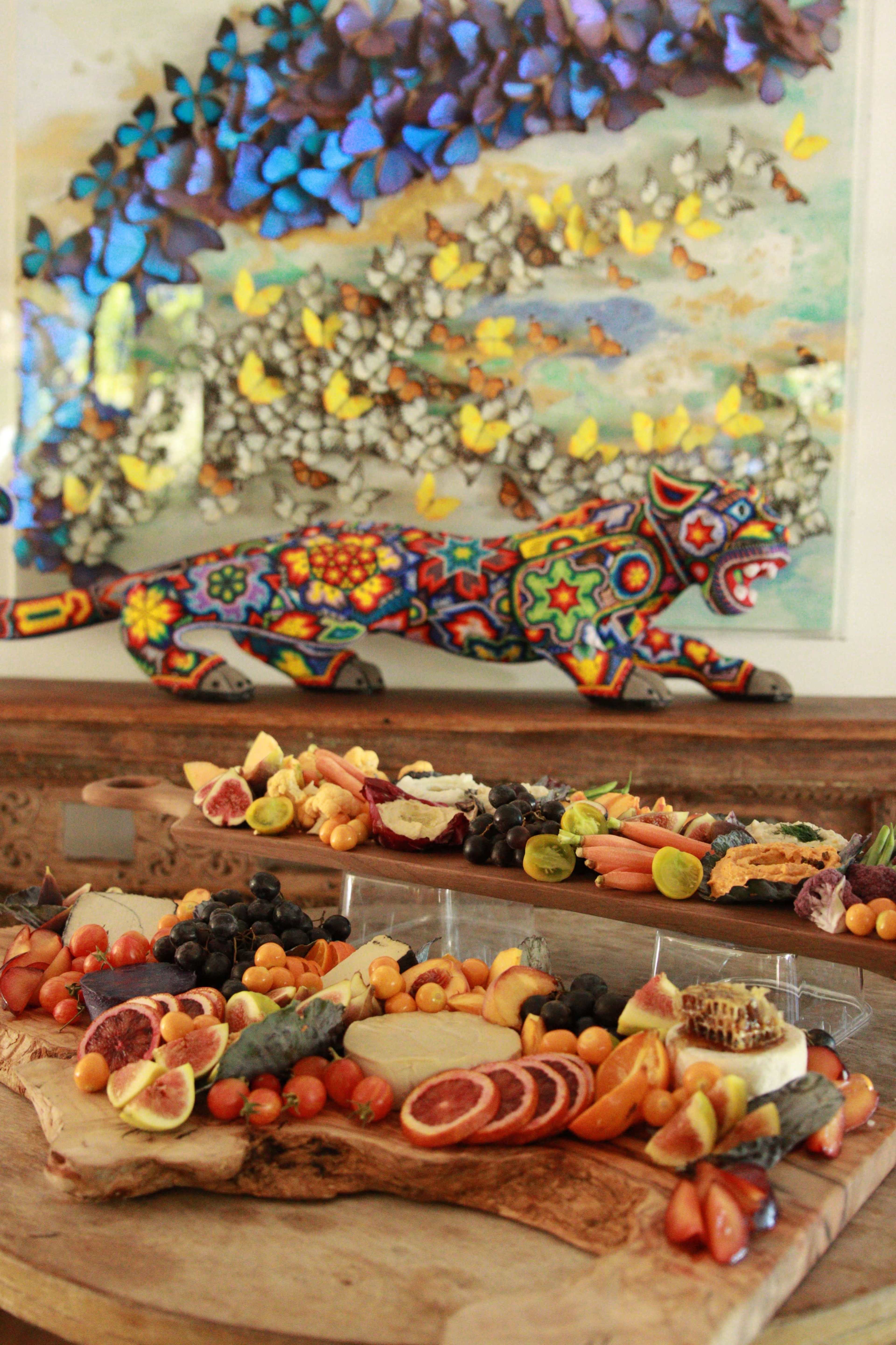 A vibrant display of assorted fruits and cheeses is arranged on a wooden board in front of a colorful, patterned sculpture of a panther and a butterfly-themed wall art.