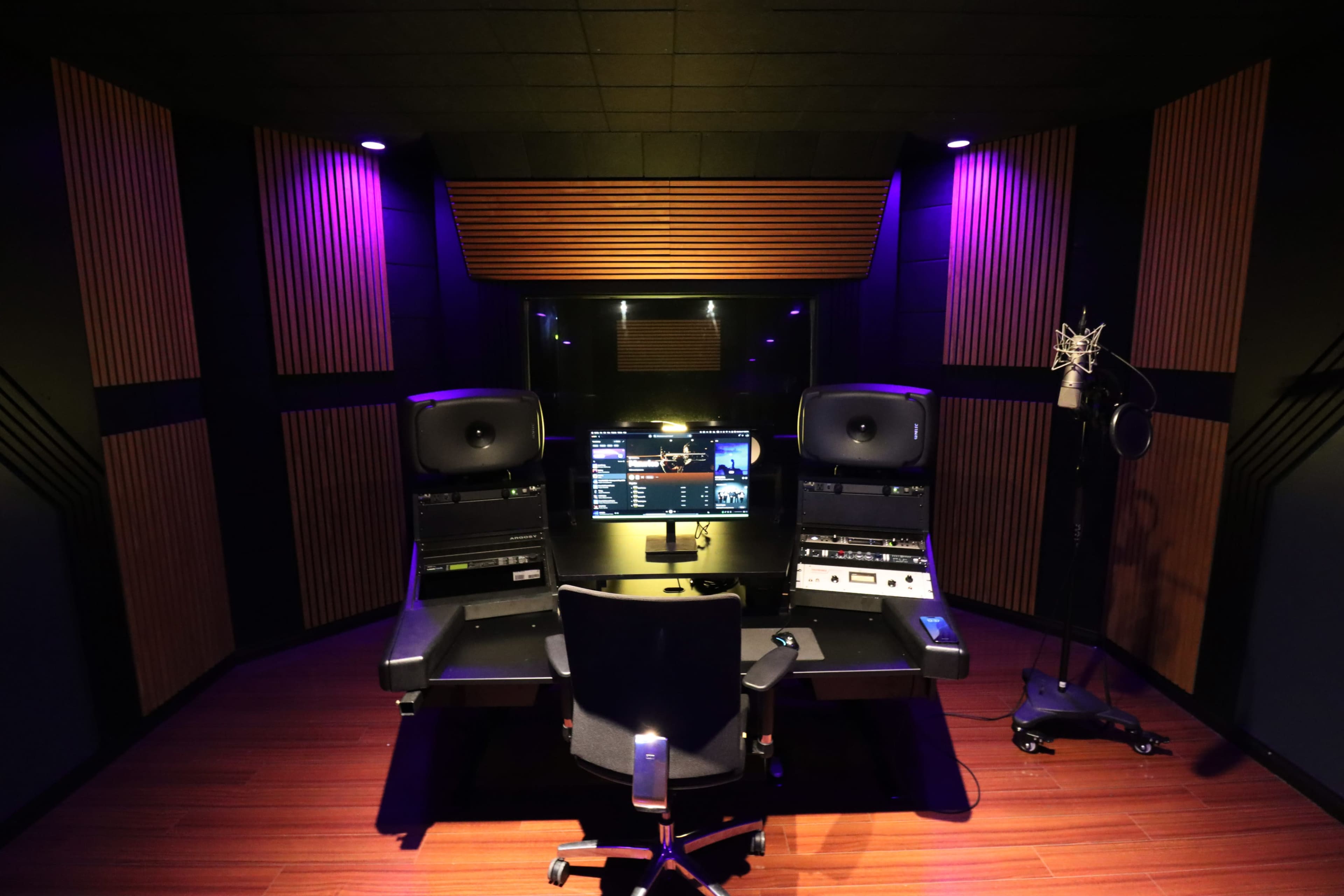 The image shows a modern recording studio featuring a central control desk with two monitor speakers, a microphone on a stand, and ambient purple lighting.