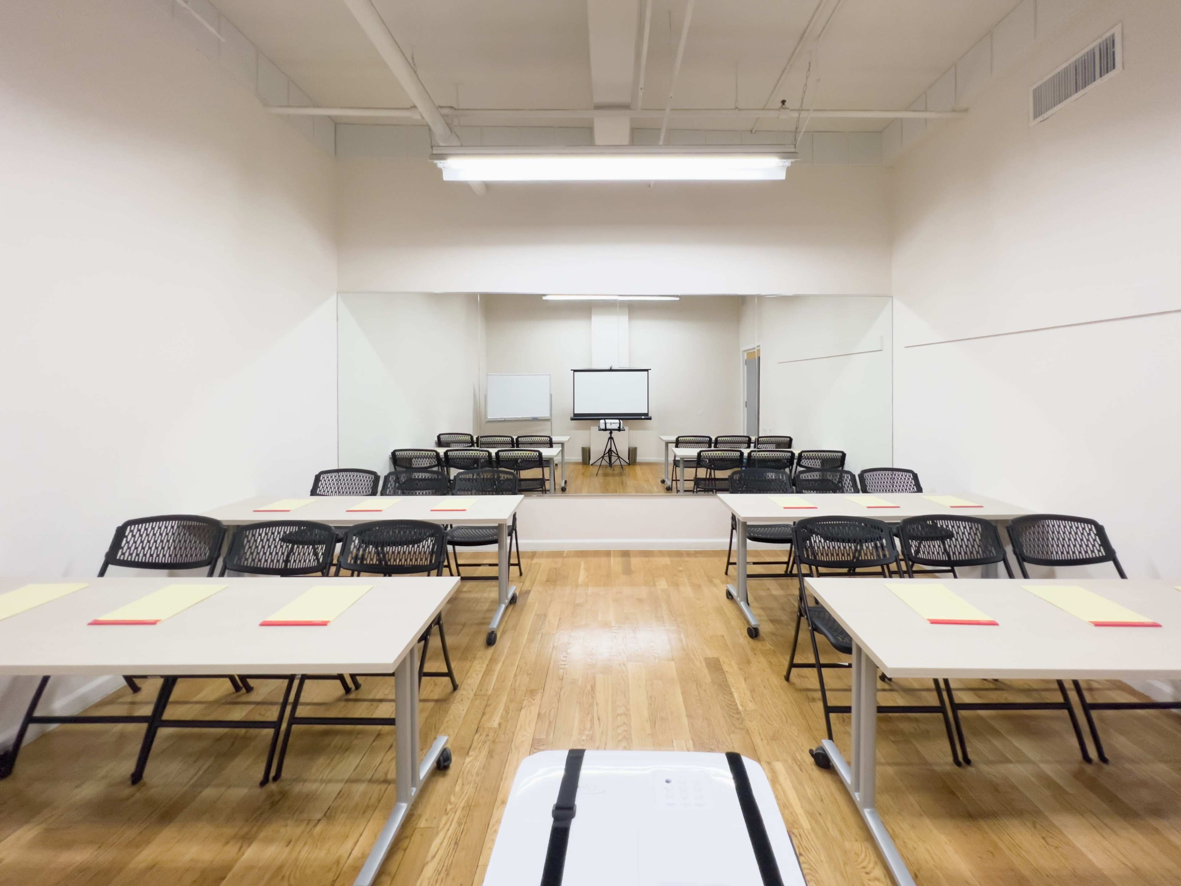 A classroom setting features multiple tables with chairs arranged facing a screen and a large mirror on one wall.