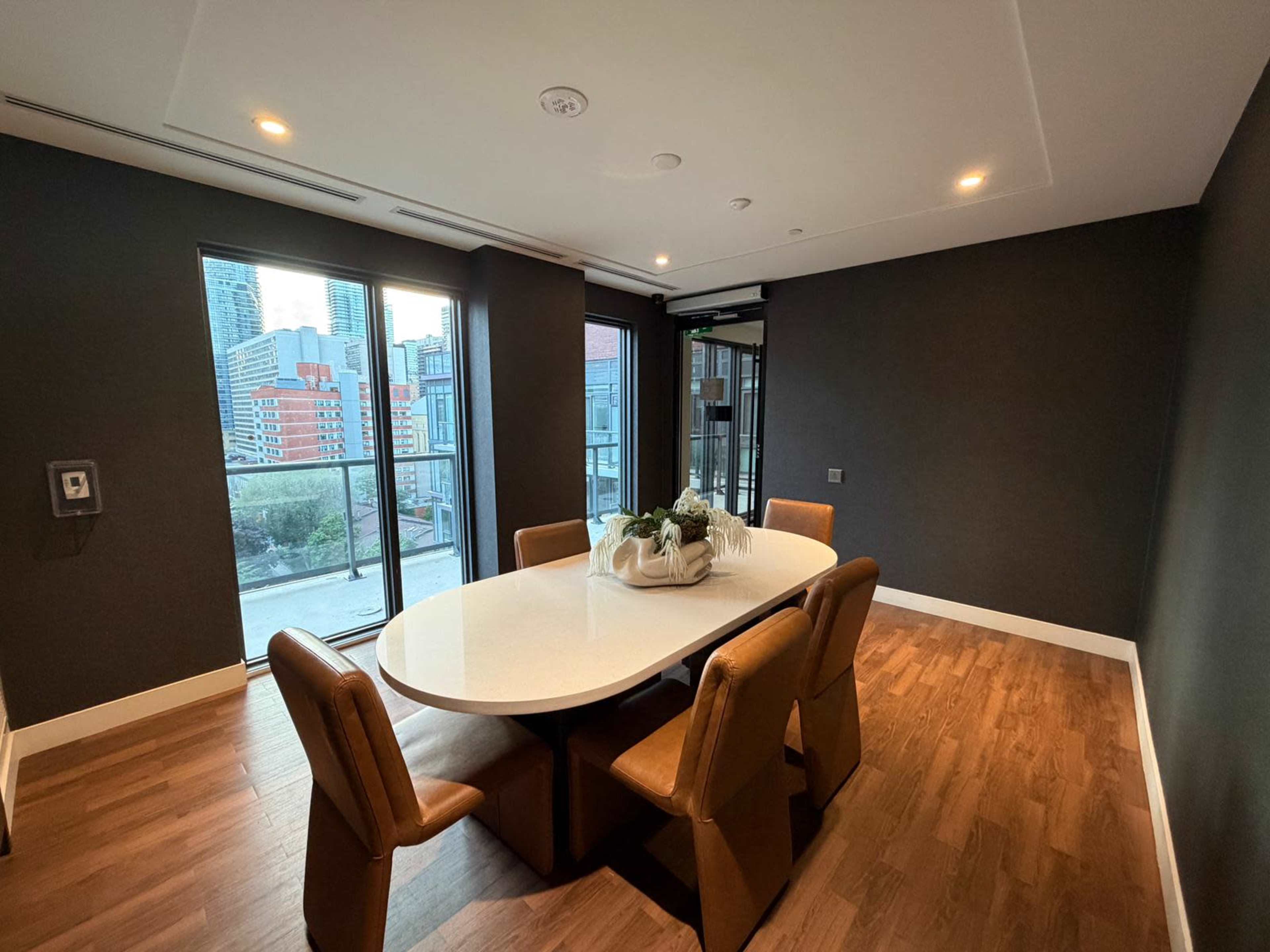 A modern dining room features a rectangular white table surrounded by brown chairs, with large windows providing a view of a cityscape.