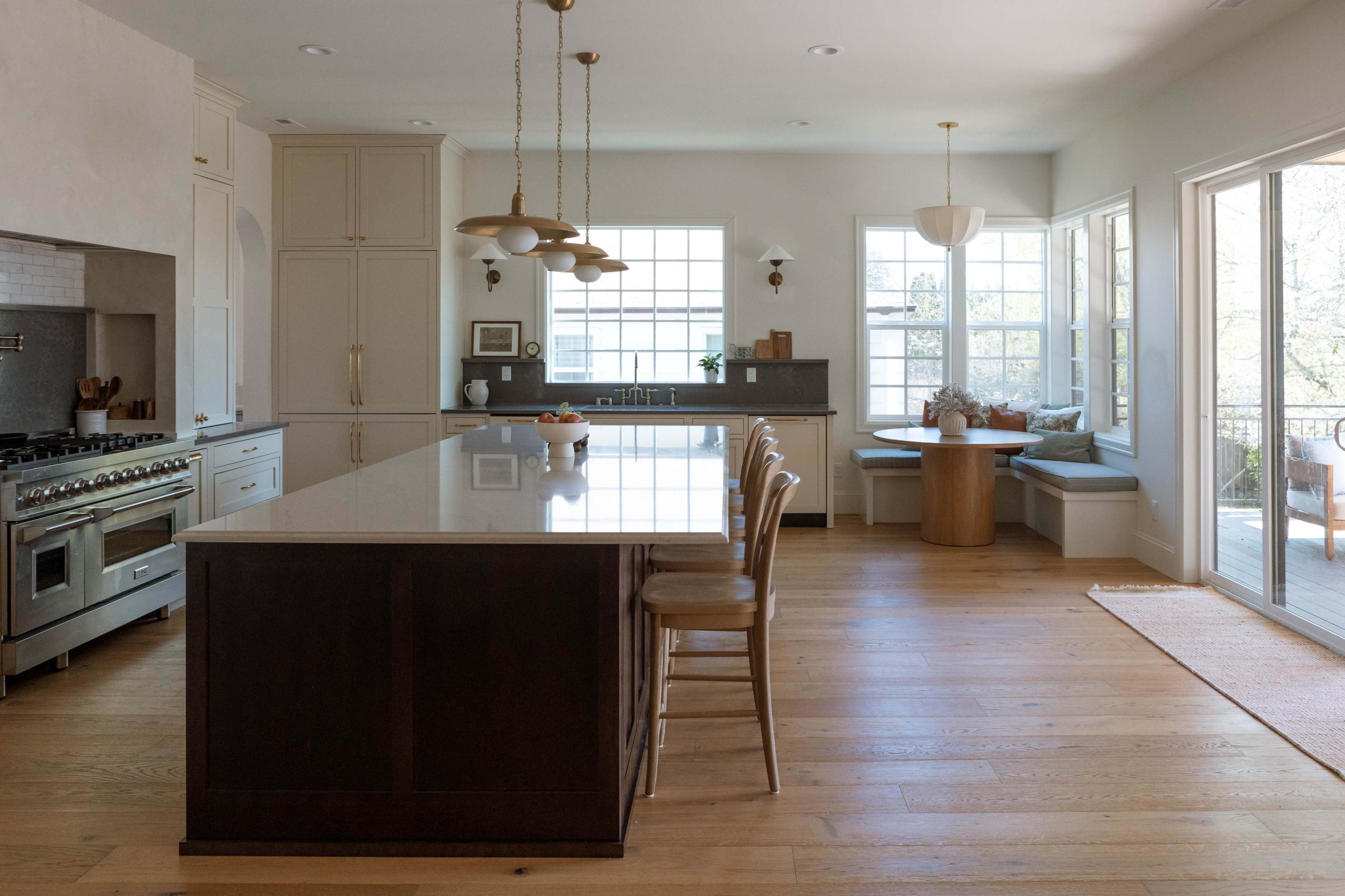 The image shows a modern kitchen featuring a large island, stainless steel appliances, and a dining area with a circular table beside windows.
