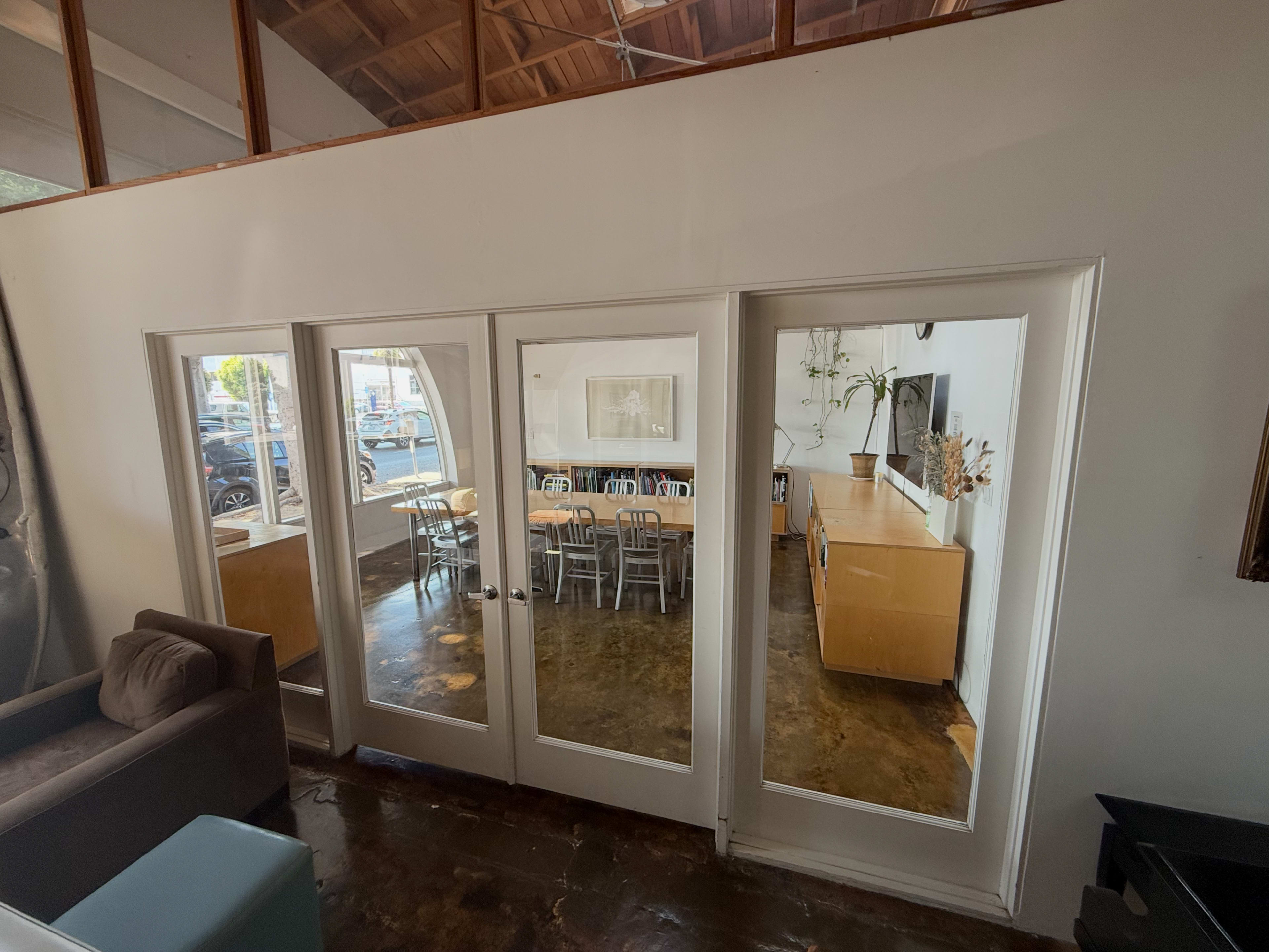 The image shows a room with a glass door entrance, leading to a dining area with a long table and chairs beside a modern kitchen.