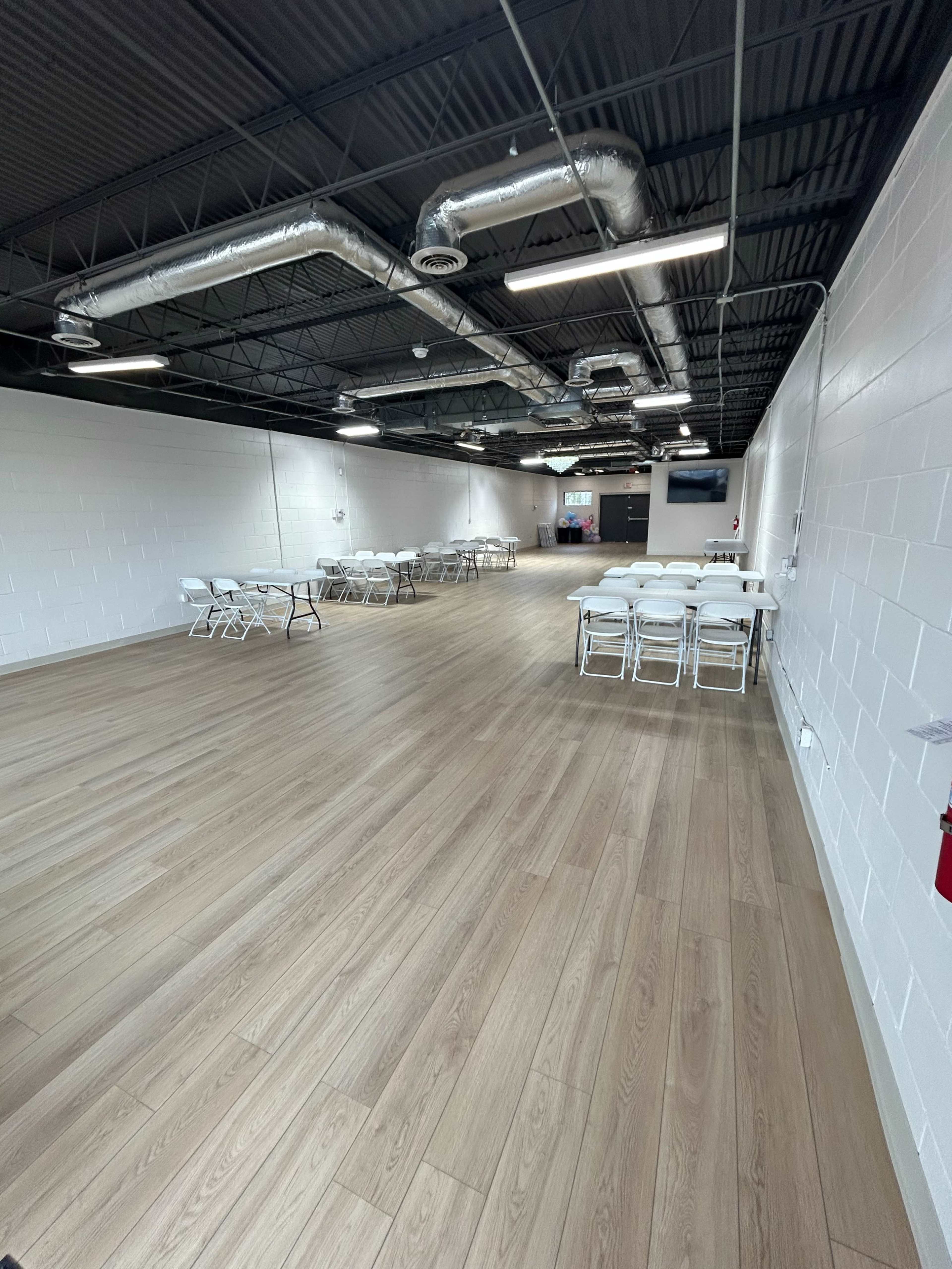 The image shows a spacious, empty room with wooden floors, white walls, and rows of folding chairs and tables arranged for an event.