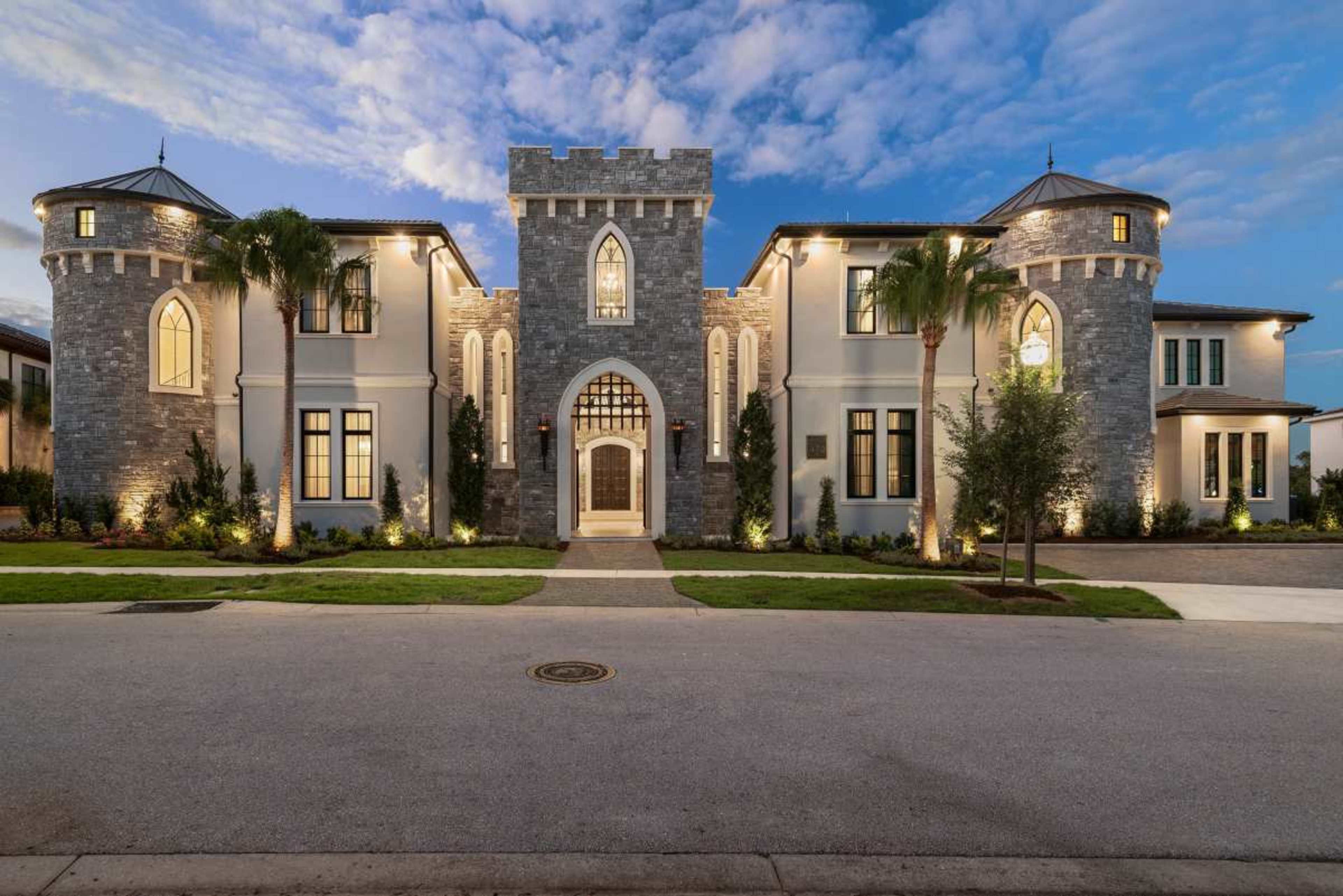 A large, ornate castle-style house features stone towers, arched windows, and a grand entrance lit by exterior lights.