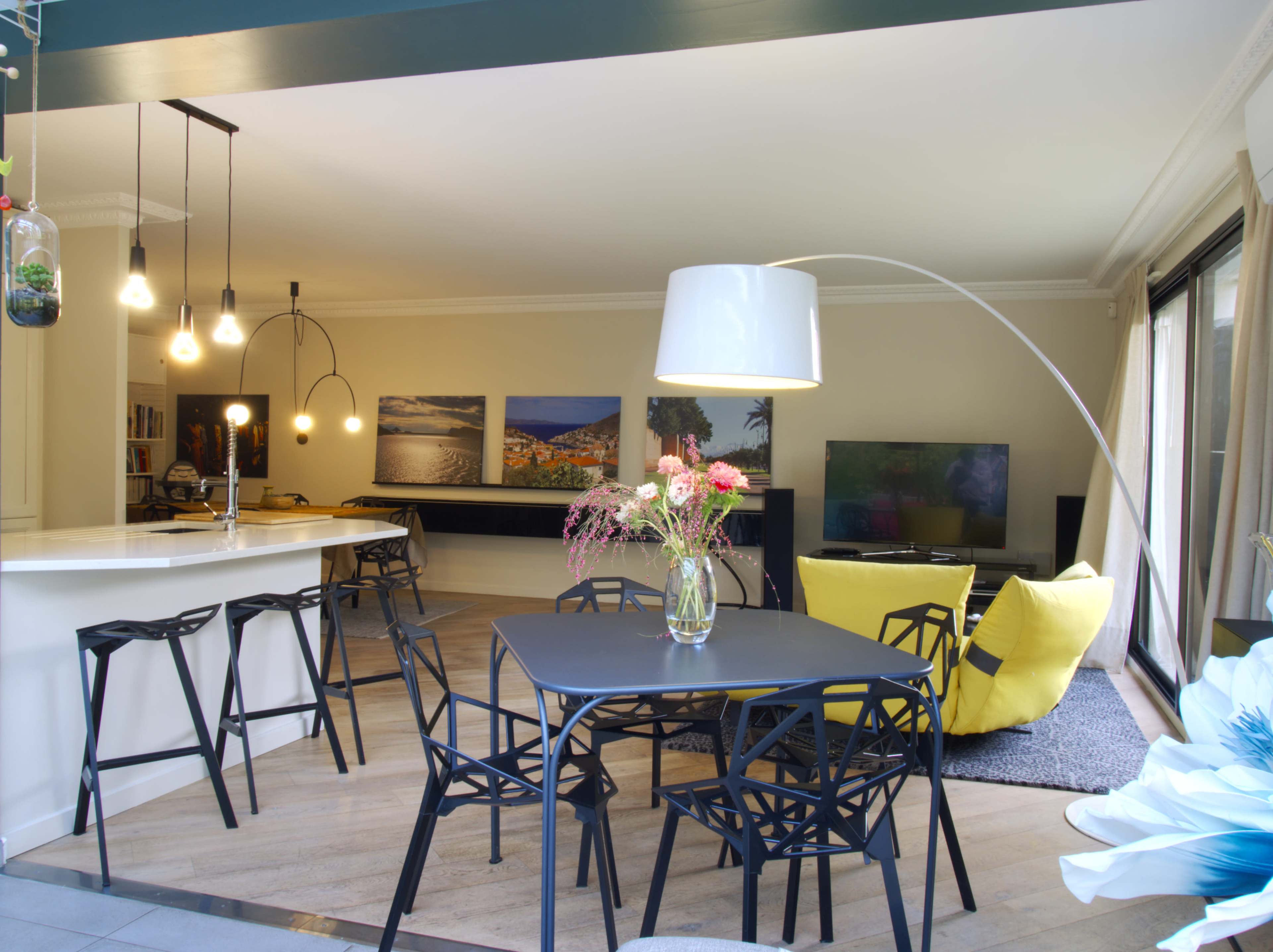 The image shows a modern, open-concept living space with a dining area featuring a black table and chairs, a curved floor lamp, and a yellow accent chair, all illuminated by pendant lights.