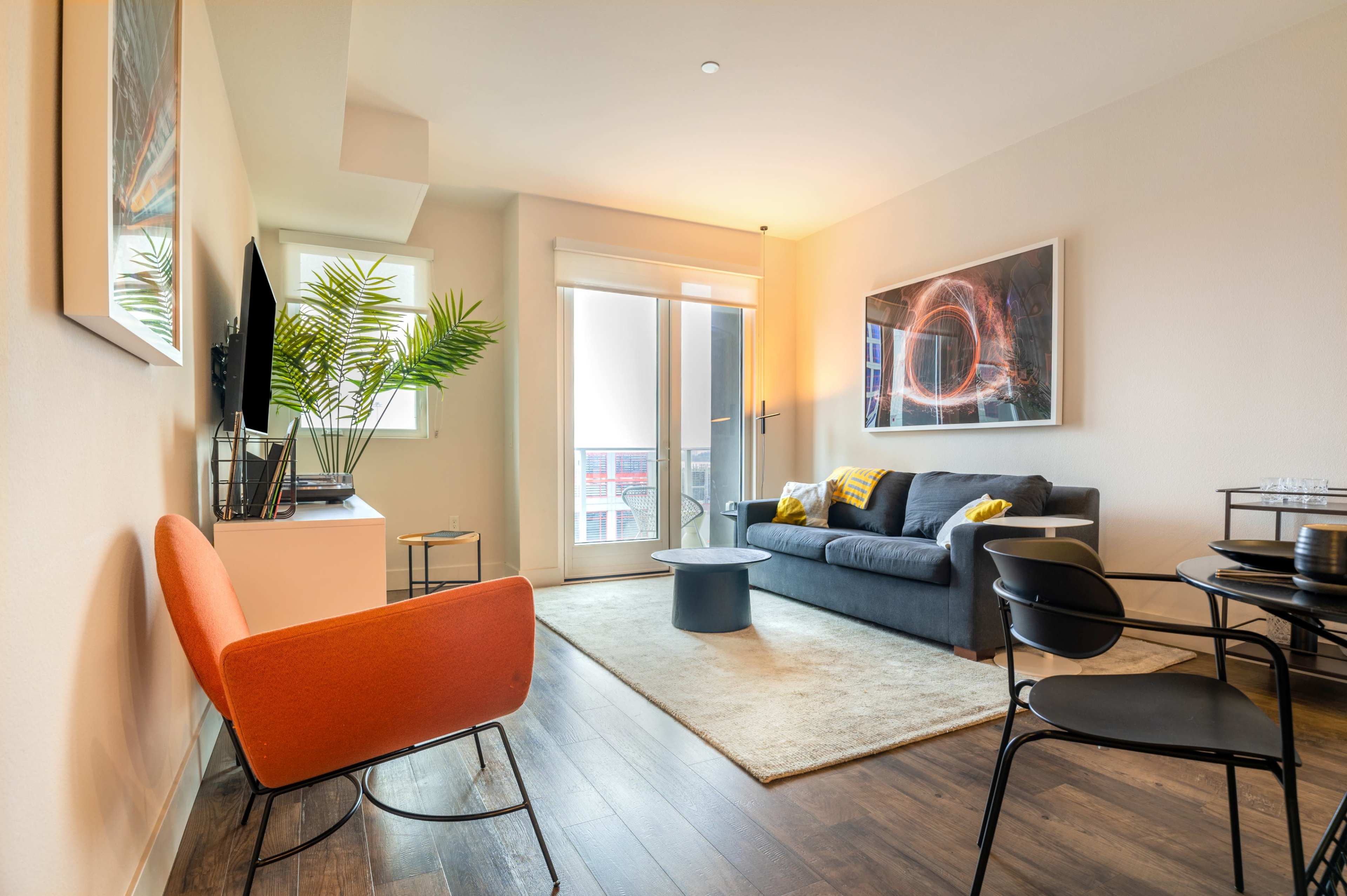 A modern living room features a gray sofa, orange chair, small black table, and large window with a view.