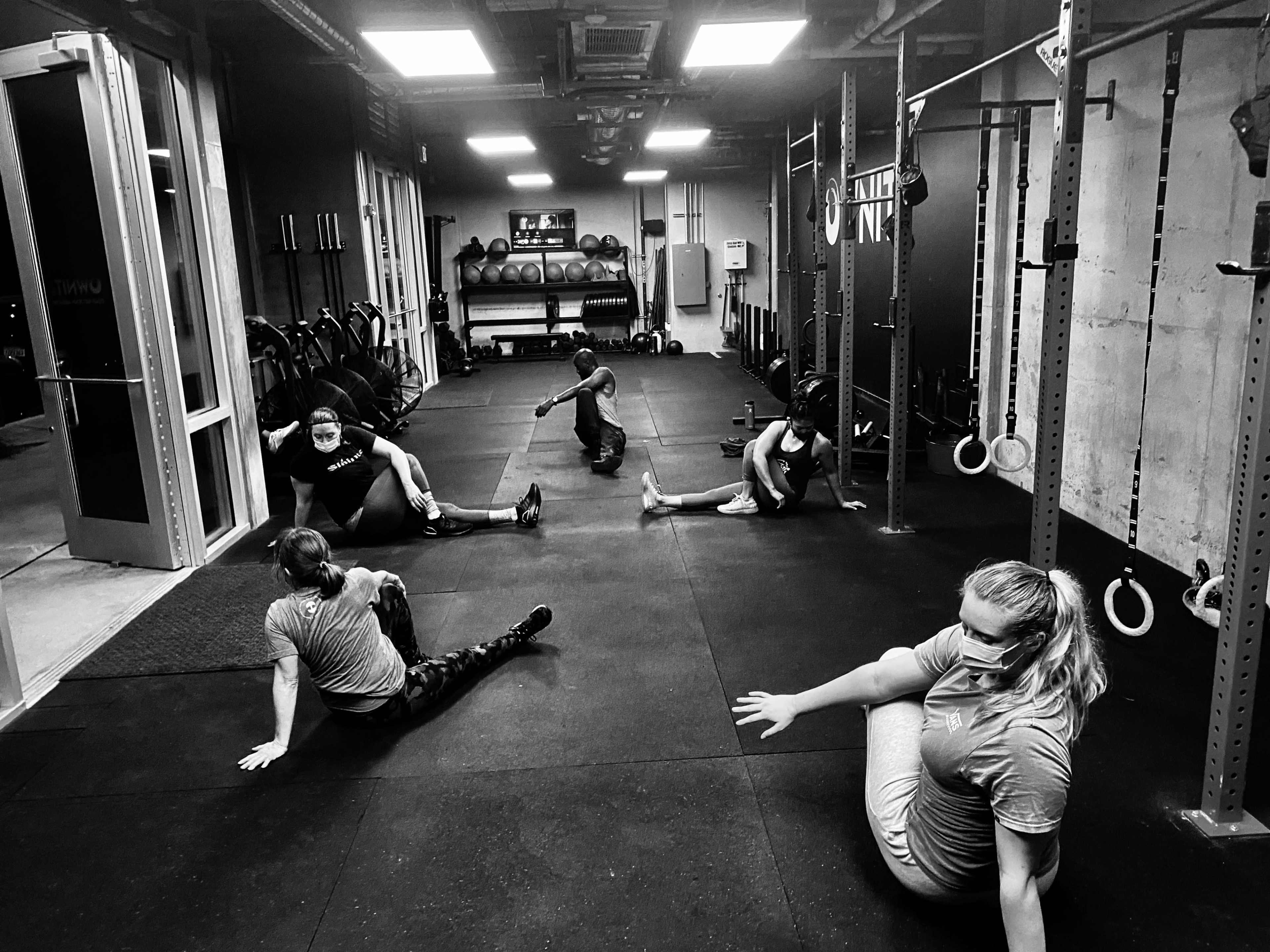 A group of individuals wearing masks is stretching on a gym floor surrounded by workout equipment.