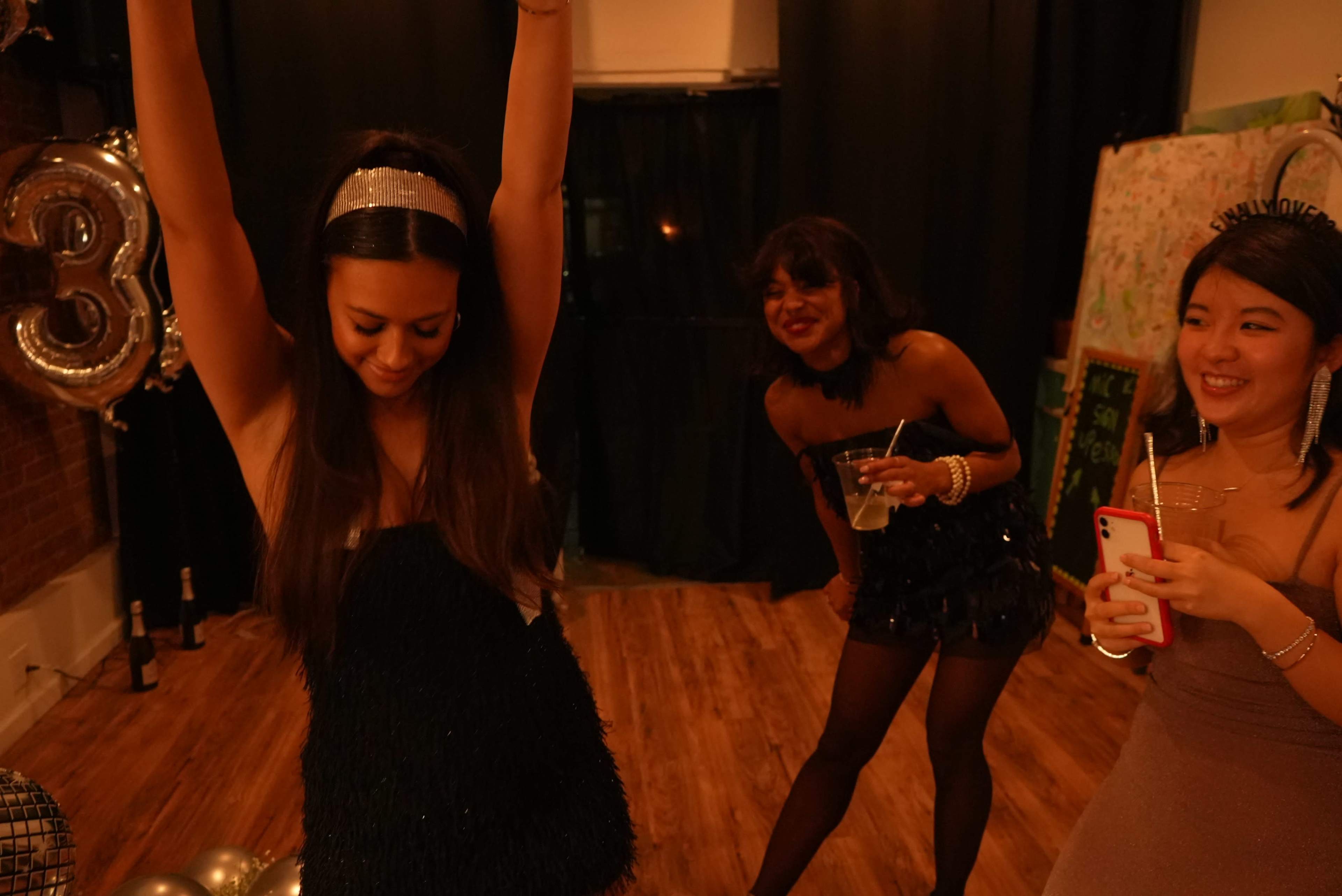 Three women are celebrating at a party, with one dancing enthusiastically while the others watch and take photos.