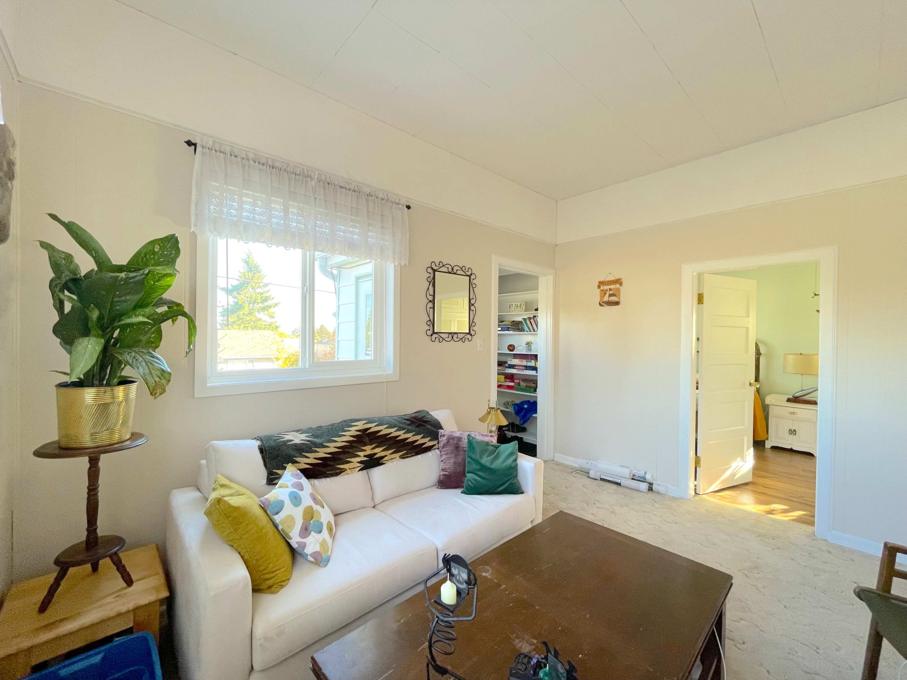 A living room with a white sofa, a wooden coffee table, a large window, and a doorway leading to another room.