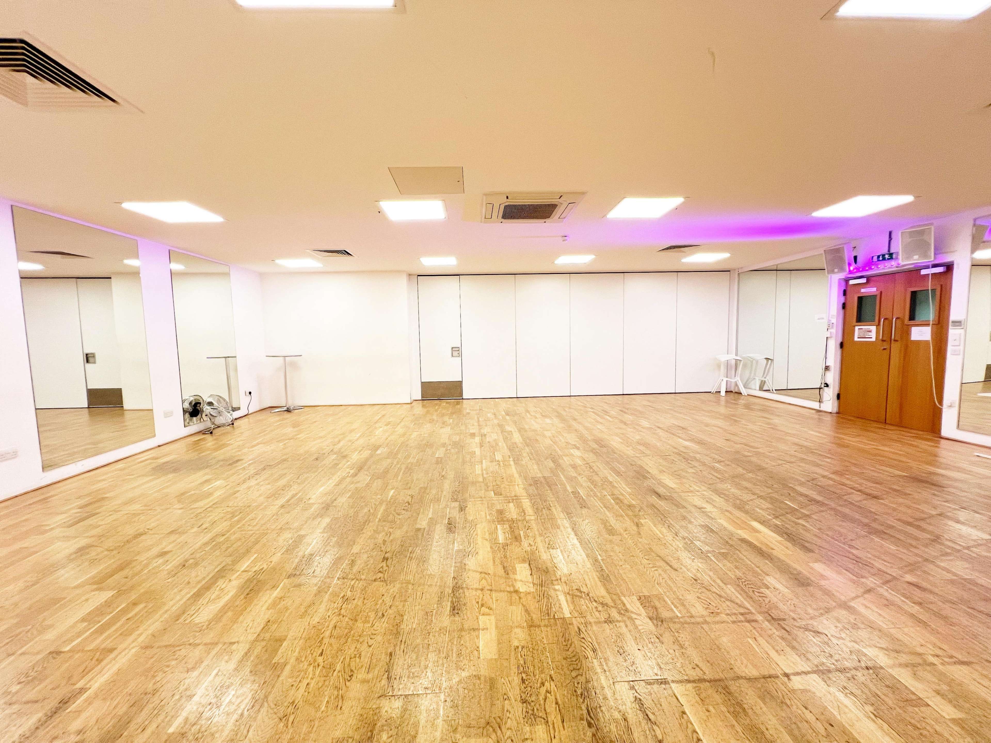 The image shows an empty, well-lit studio space with wooden flooring, large mirrors on one wall, and a white sliding door.