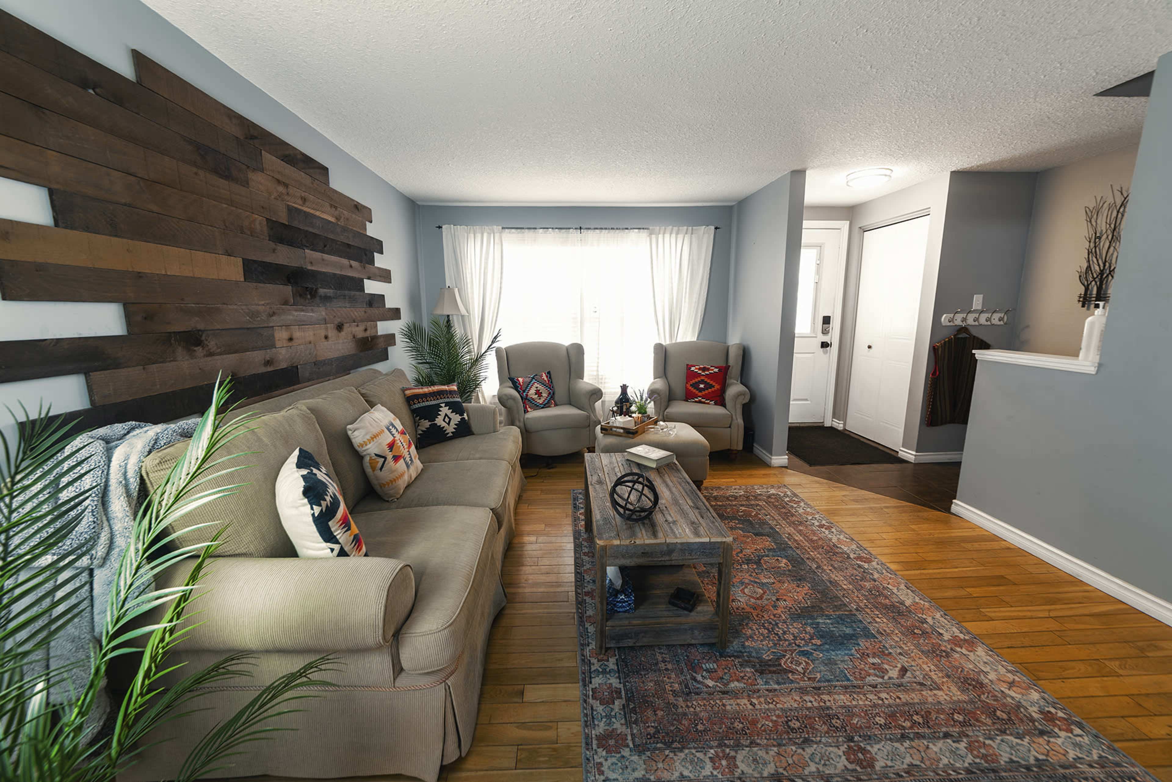 A living room features a wooden accent wall, a beige couch with decorative pillows, two armchairs, a coffee table, and a patterned area rug.