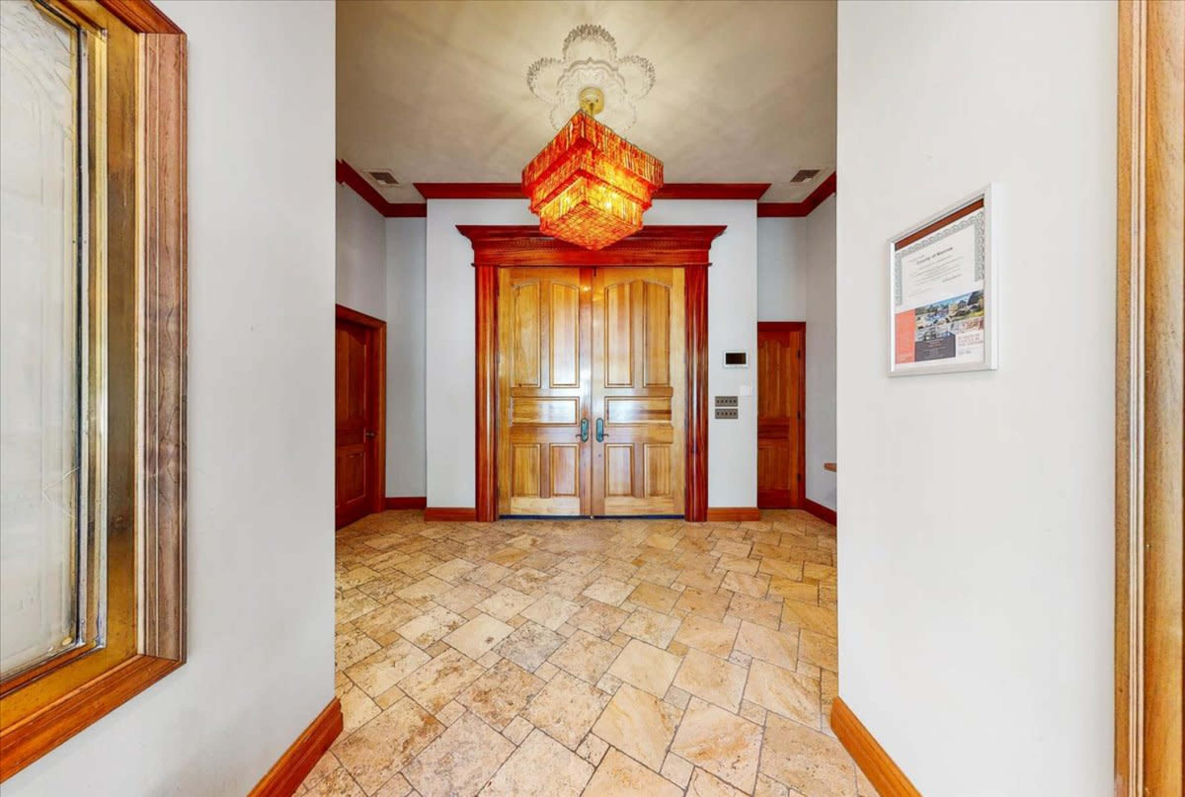 A hallway leading to large wooden double doors, with a chandelier hanging above and tiled flooring.