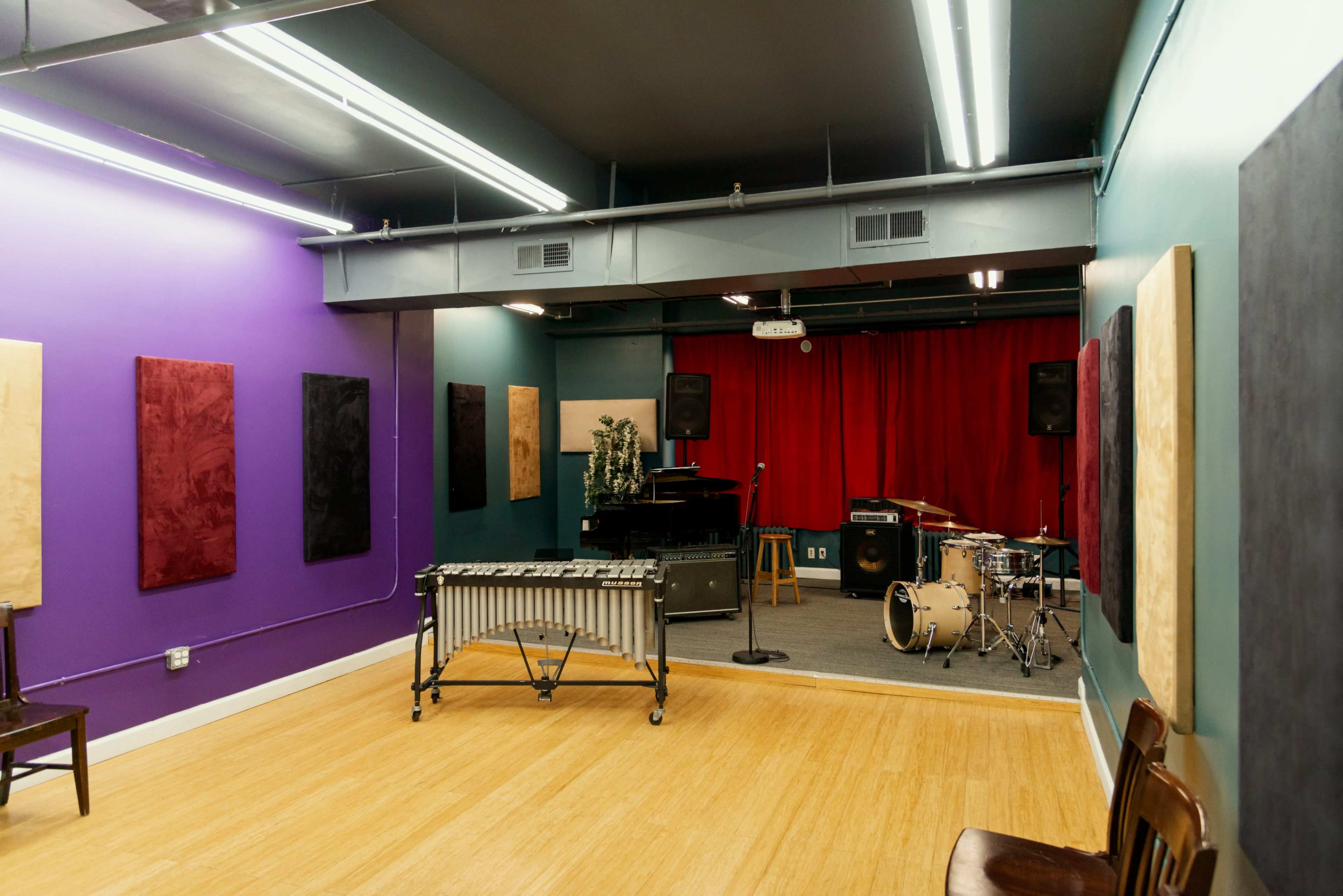 The image shows a colorful music rehearsal space featuring a drum set, a vibraphone on wheels, and a small stage with red curtains.
