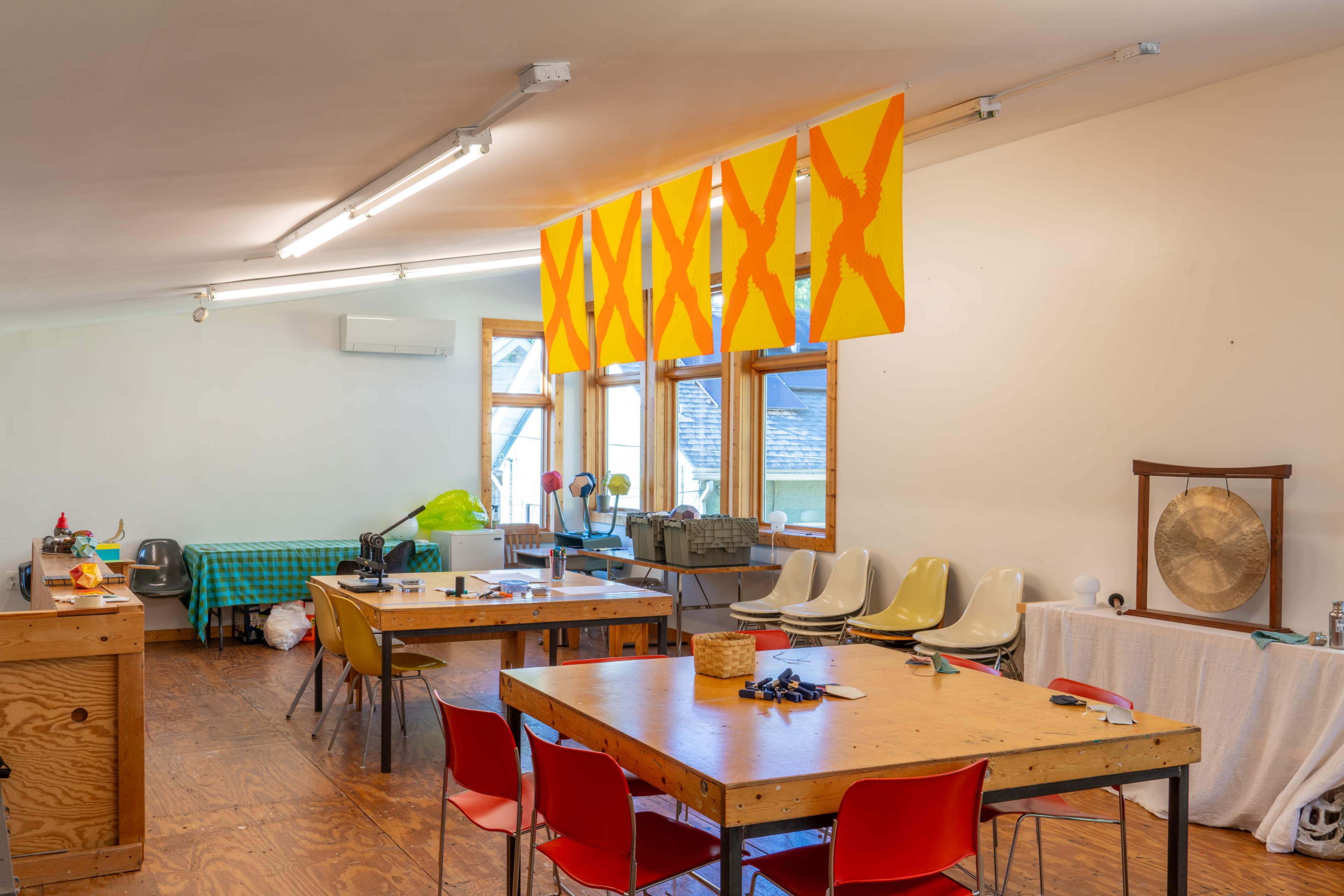 The image shows a bright, spacious workshop with several wooden tables, colorful chairs, and large orange and yellow artwork displayed on the ceiling.
