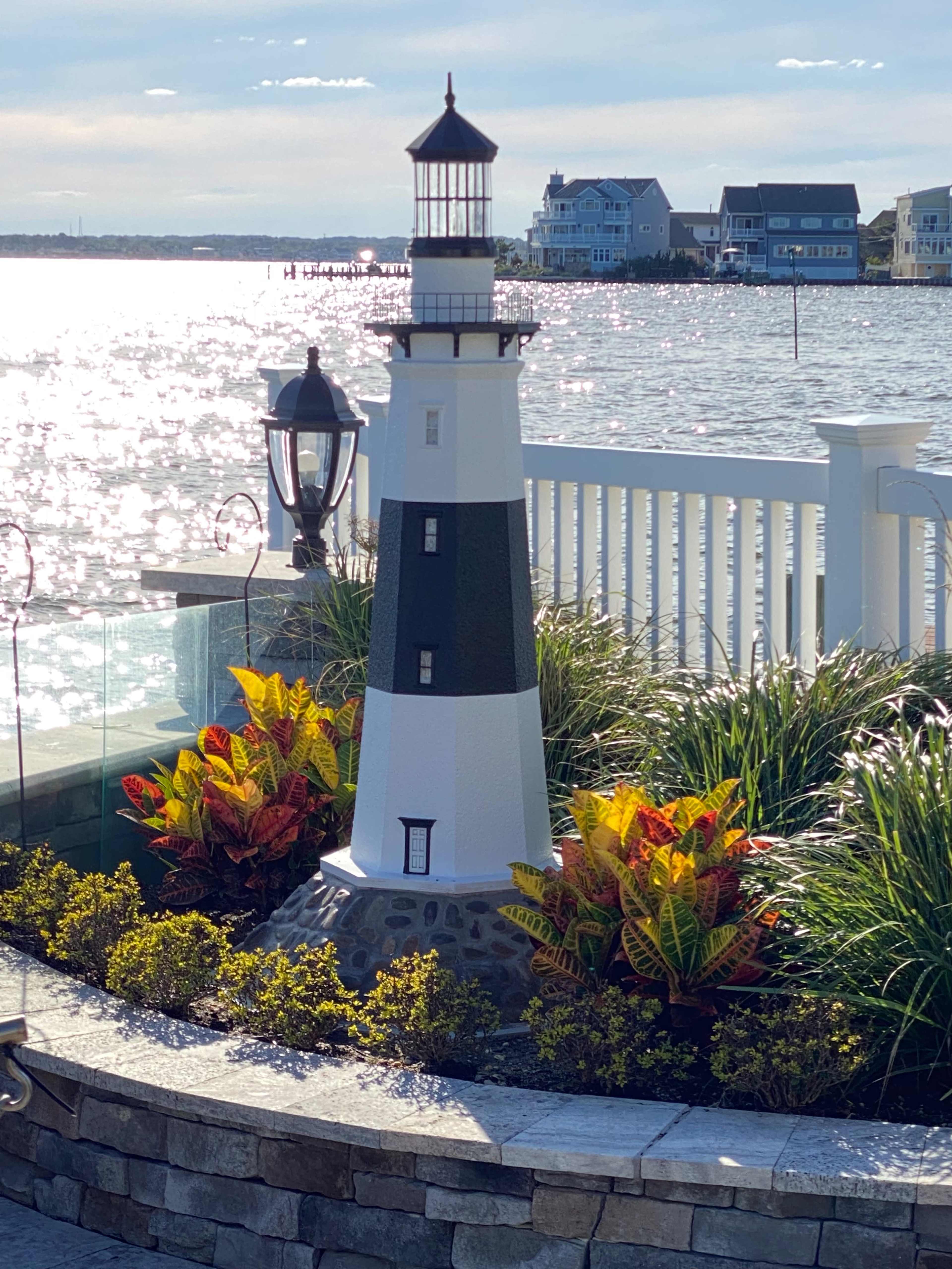 A small lighthouse sculpture surrounded by colorful plants sits on a stone terrace overlooking a shimmering body of water.