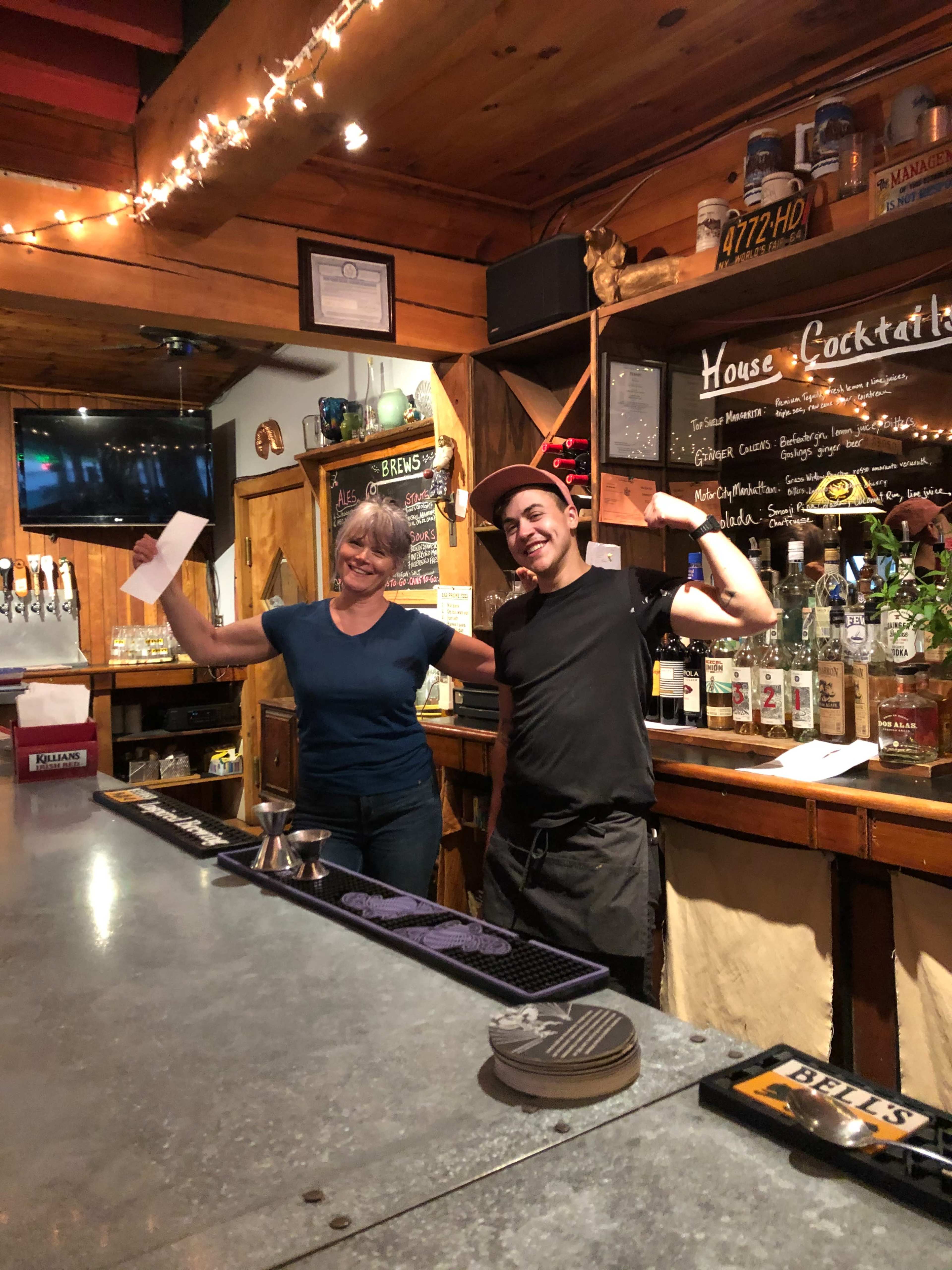 A man and a woman smile and wave behind a bar filled with various bottles and a beer tap setup.