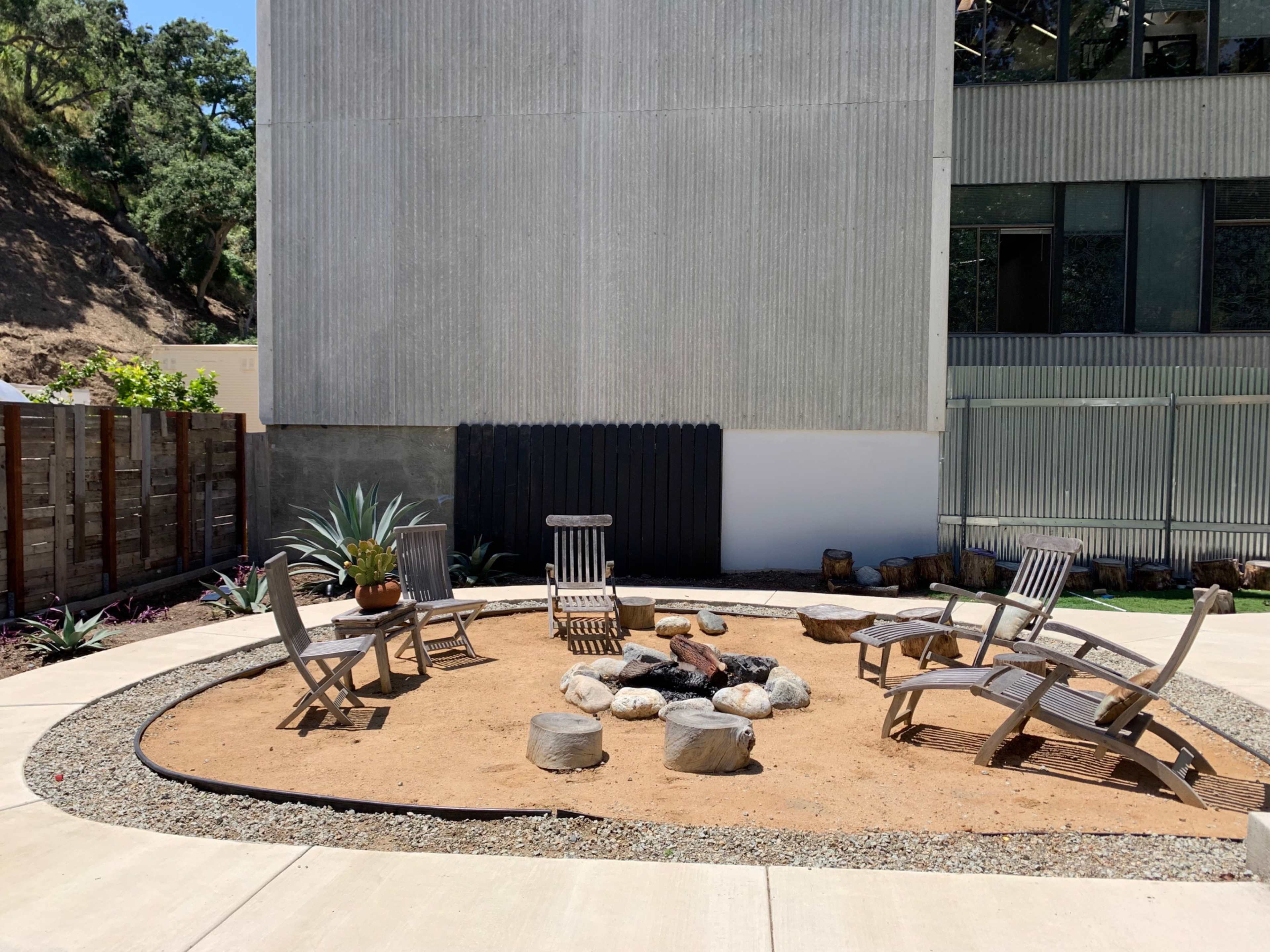 The image shows a circular seating area with wooden chairs arranged around a fire pit surrounded by gravel and rocks, located next to a modern building.