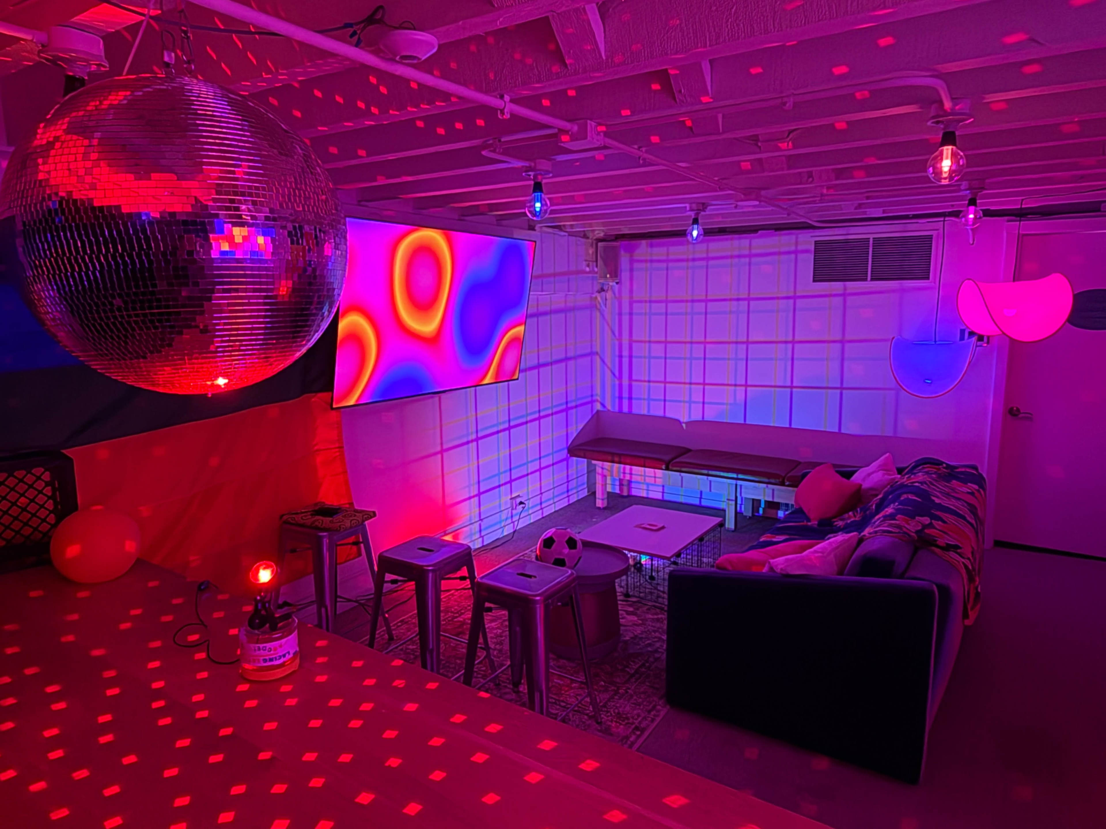 The image shows a modern lounge area with colorful lighting, including a disco ball and a large screen displaying abstract patterns, along with a sofa, tables, and decorative items.
