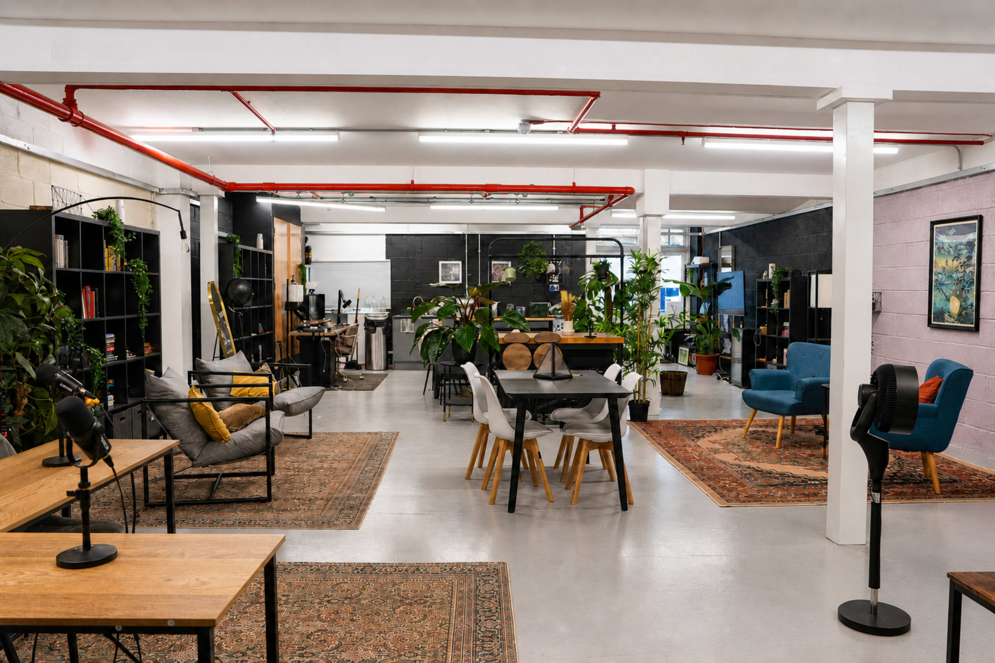 The image shows a spacious office interior featuring wooden tables, chairs, plants, and various workstations with shelving and artwork on the walls.