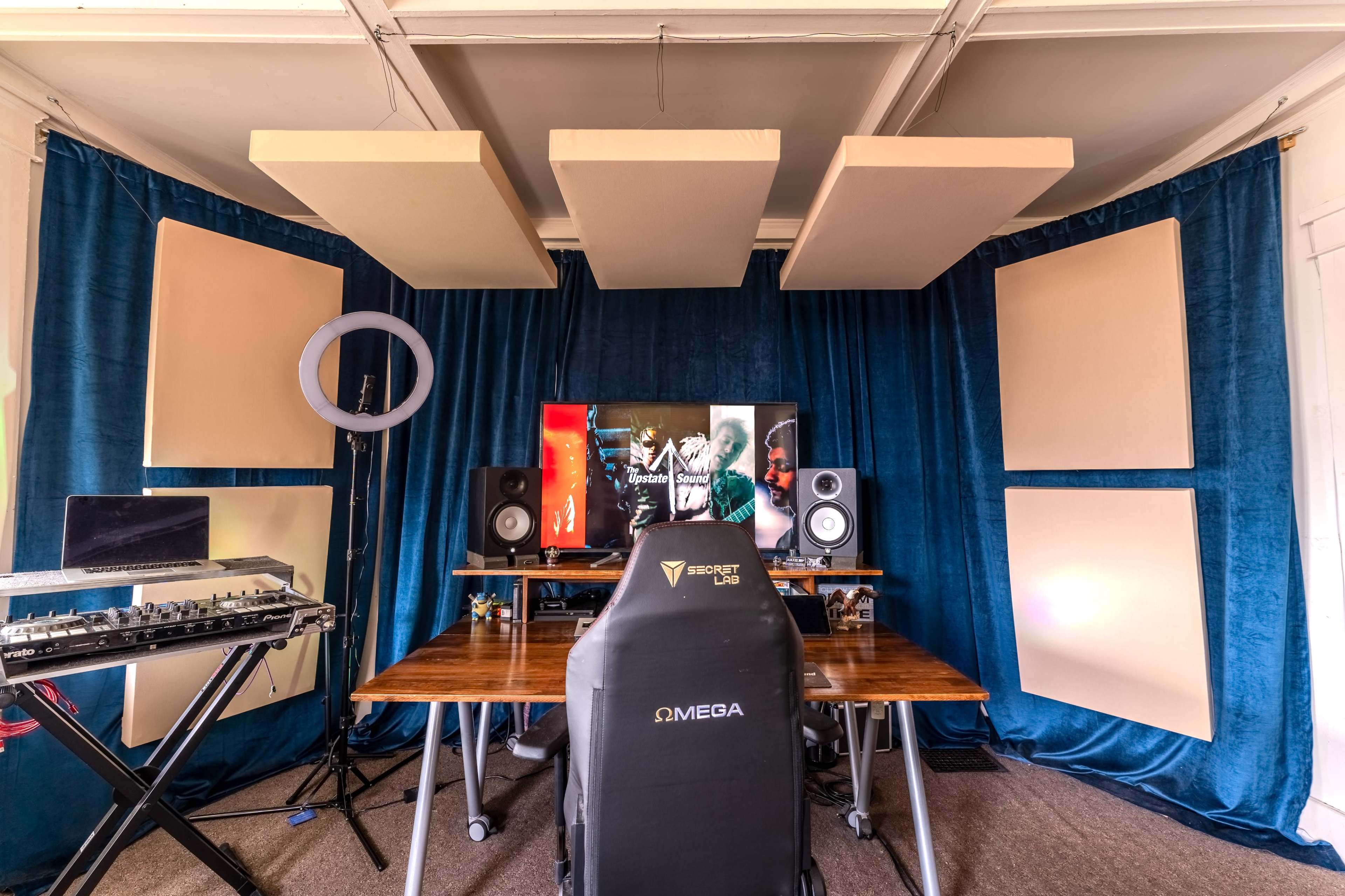 A music production setup featuring a desk with a keyboard, speakers, and a large monitor surrounded by acoustic panels and blue curtains.