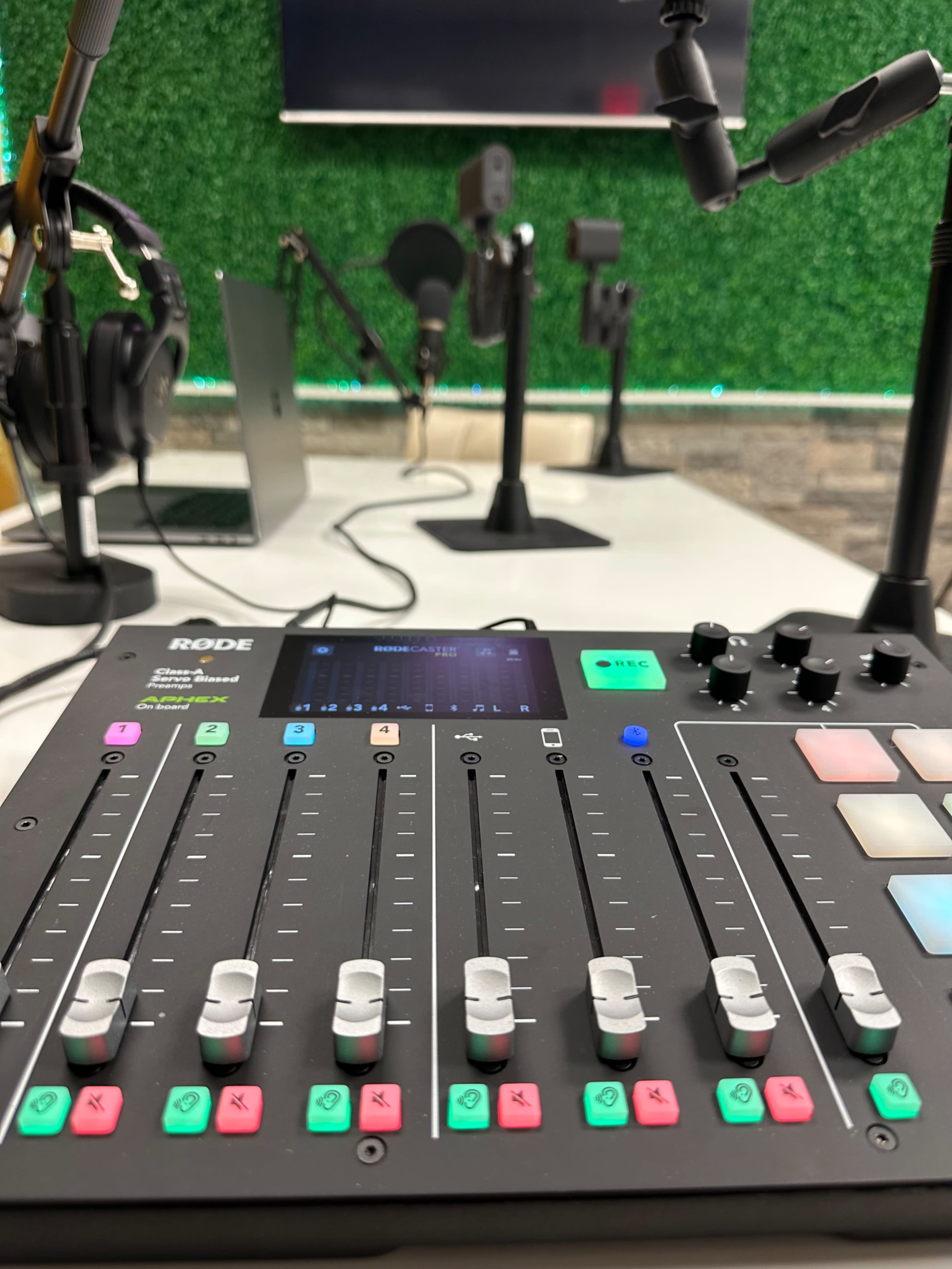 A close-up view of a Rode audio mixer with sliders and buttons, placed on a table in a recording studio featuring microphones and headphones in the background.